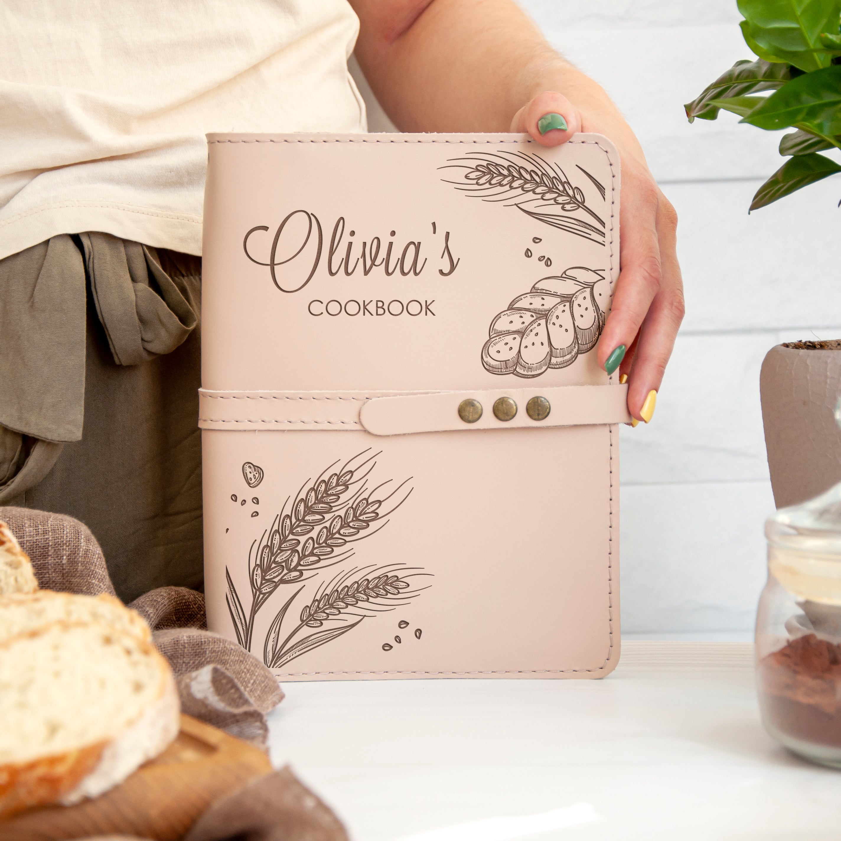 A person is holding a personalized cookbook with a design of wheat and bread on the cover, which is placed on a table with various food items and a potted plant in the background.