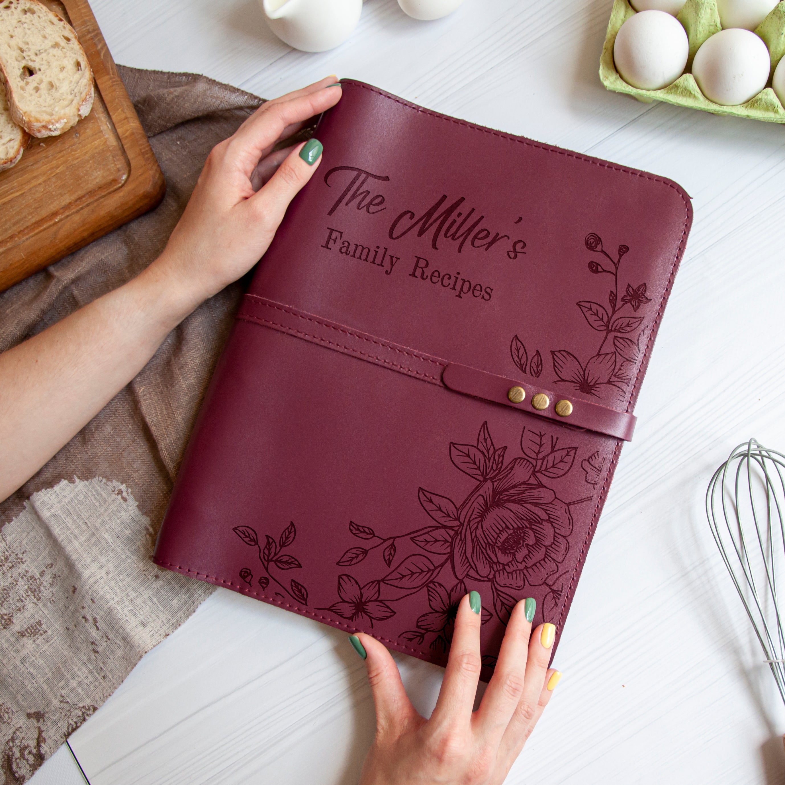 A person's hands holding a burgundy leather recipe book with floral designs, placed on a white surface next to a wooden cutting board, a whisk, and a carton of eggs.