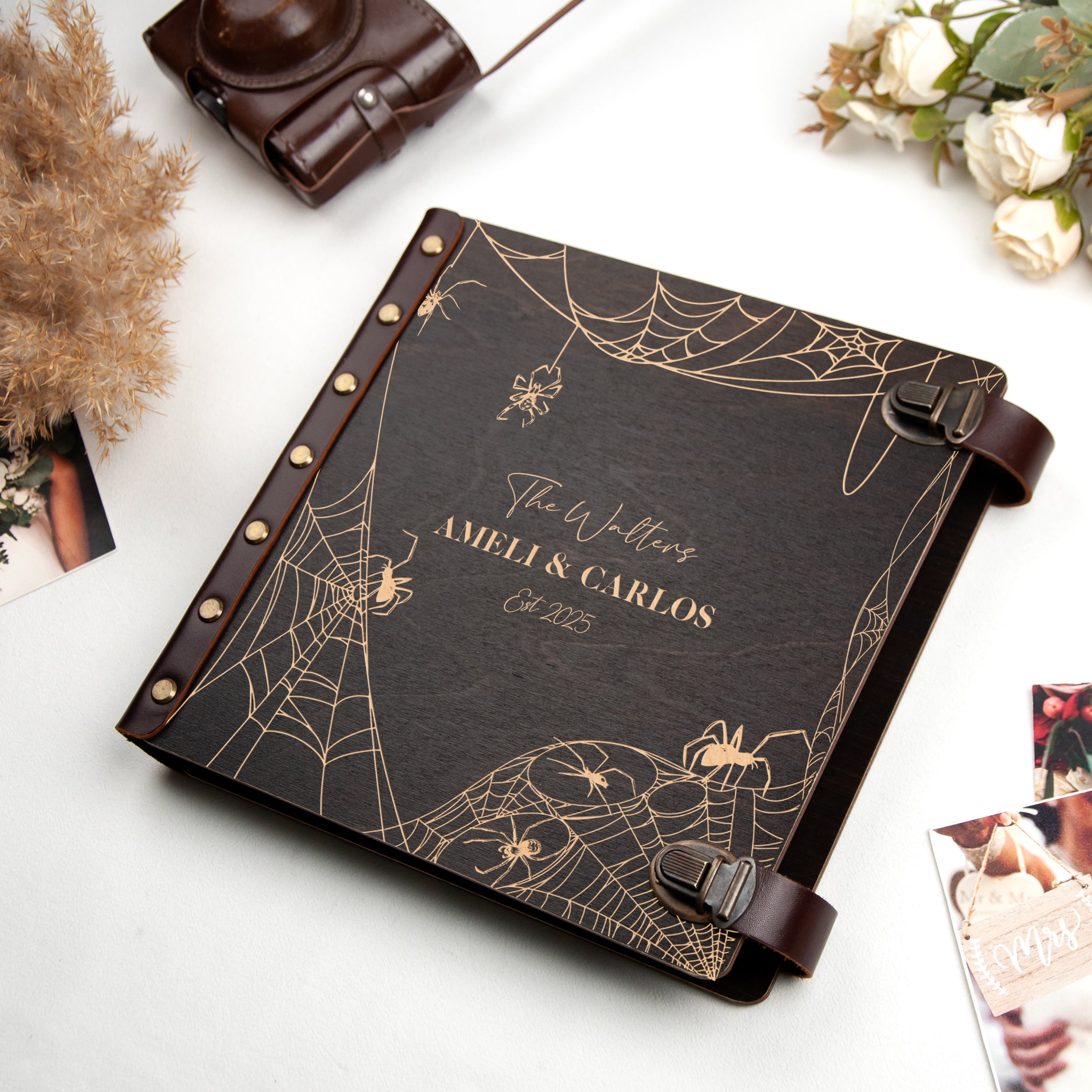 A black leather-bound book titled "The Wedding" by Ameli & Carlos, featuring a spider web design, sits on a table surrounded by other items such as a camera, flowers, and photographs.