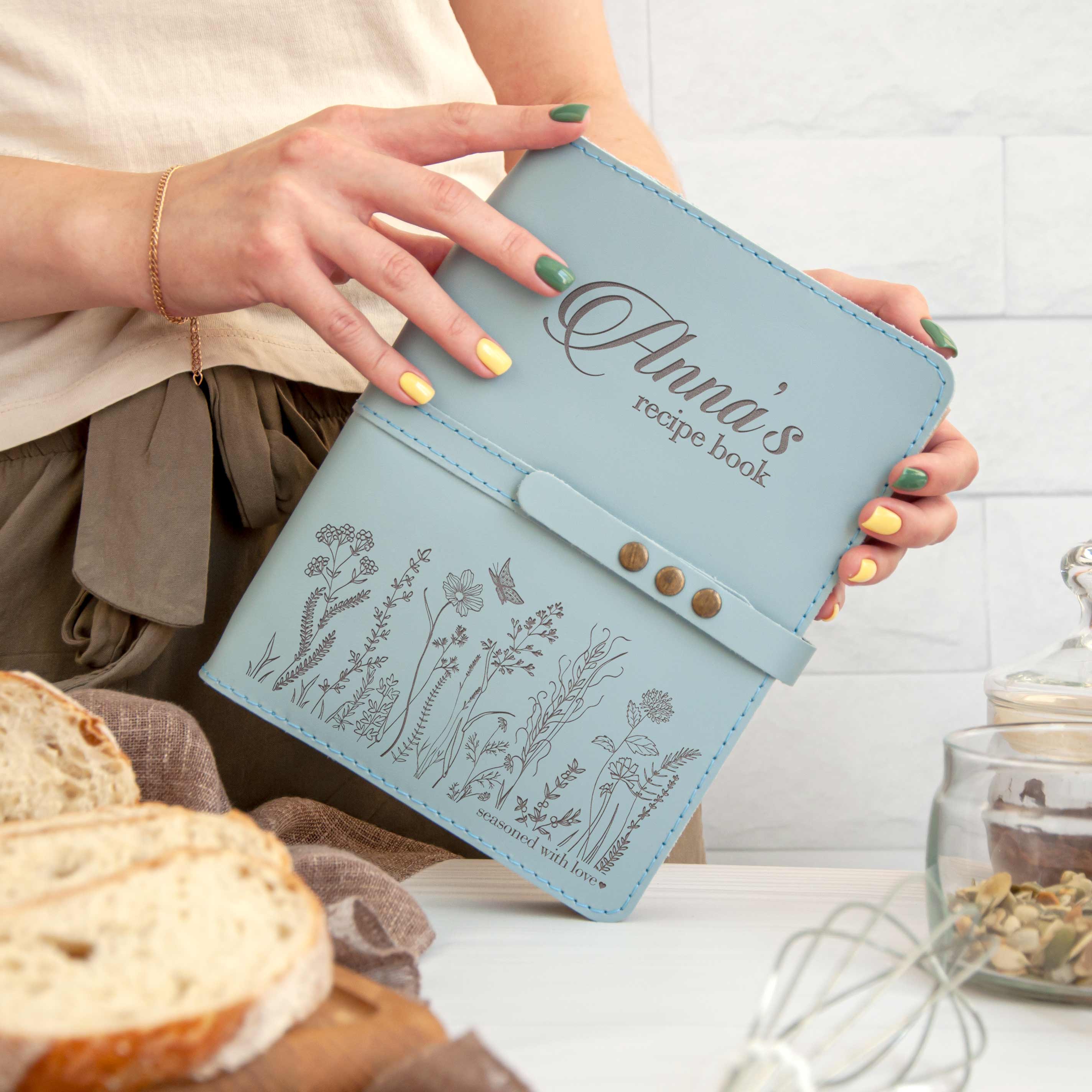 A person holding a light blue recipe book with floral designs, standing in front of a table with bread and other food items.