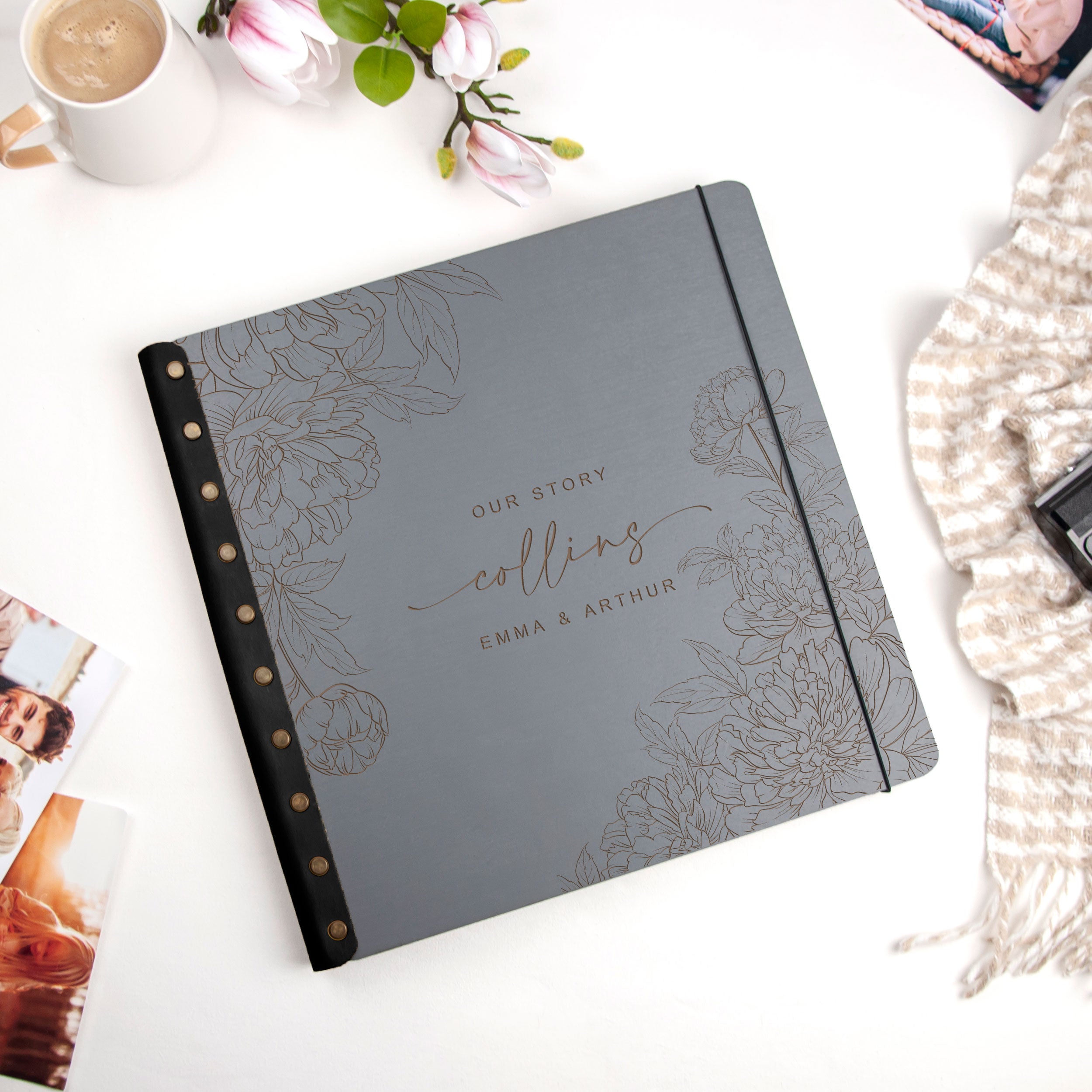 A gray notebook with a floral design and the text "Our Story Collins" printed on it, placed on a white surface alongside other items such as a coffee cup, flowers, and a scarf.