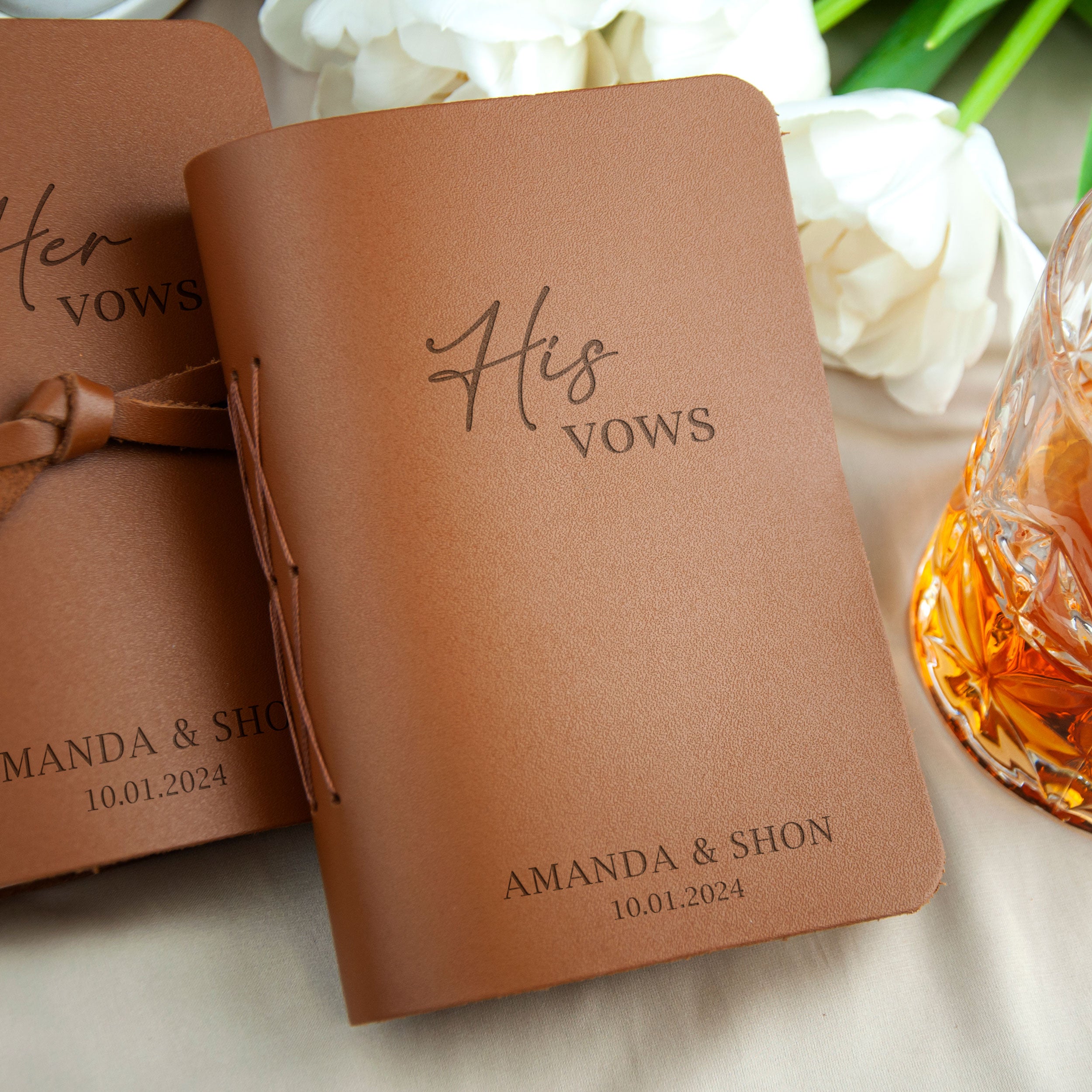 two leather-bound books with the text "His Vows" printed on them, placed on a table with a glass of amber liquid and white flowers in the background.