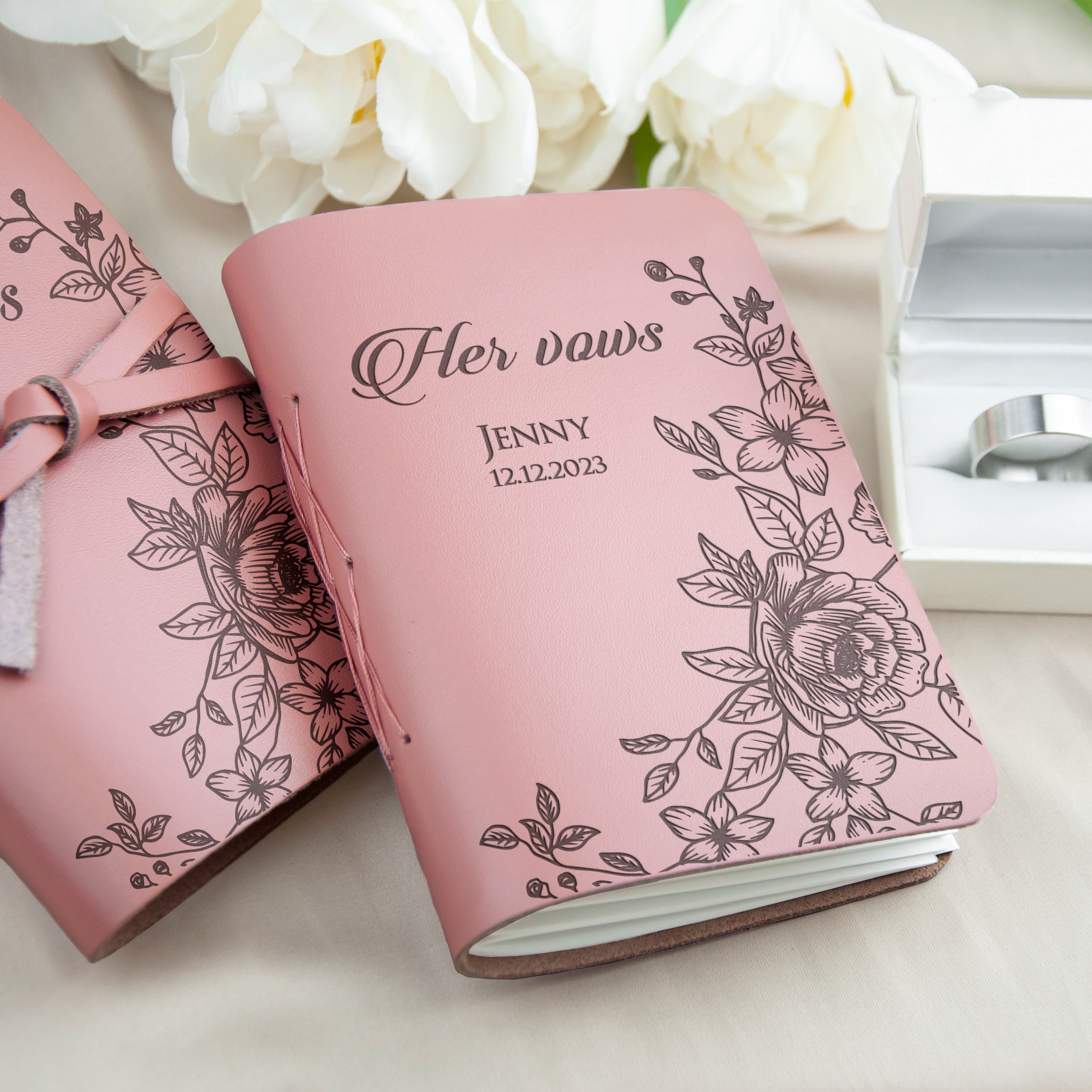 A pink leather-bound book with a floral design and the text "Her vows" printed on it, along with the date "Jenny 12.12.2023" printed below. The book is placed next to a white box containing a ring, and there are white flowers in the background.