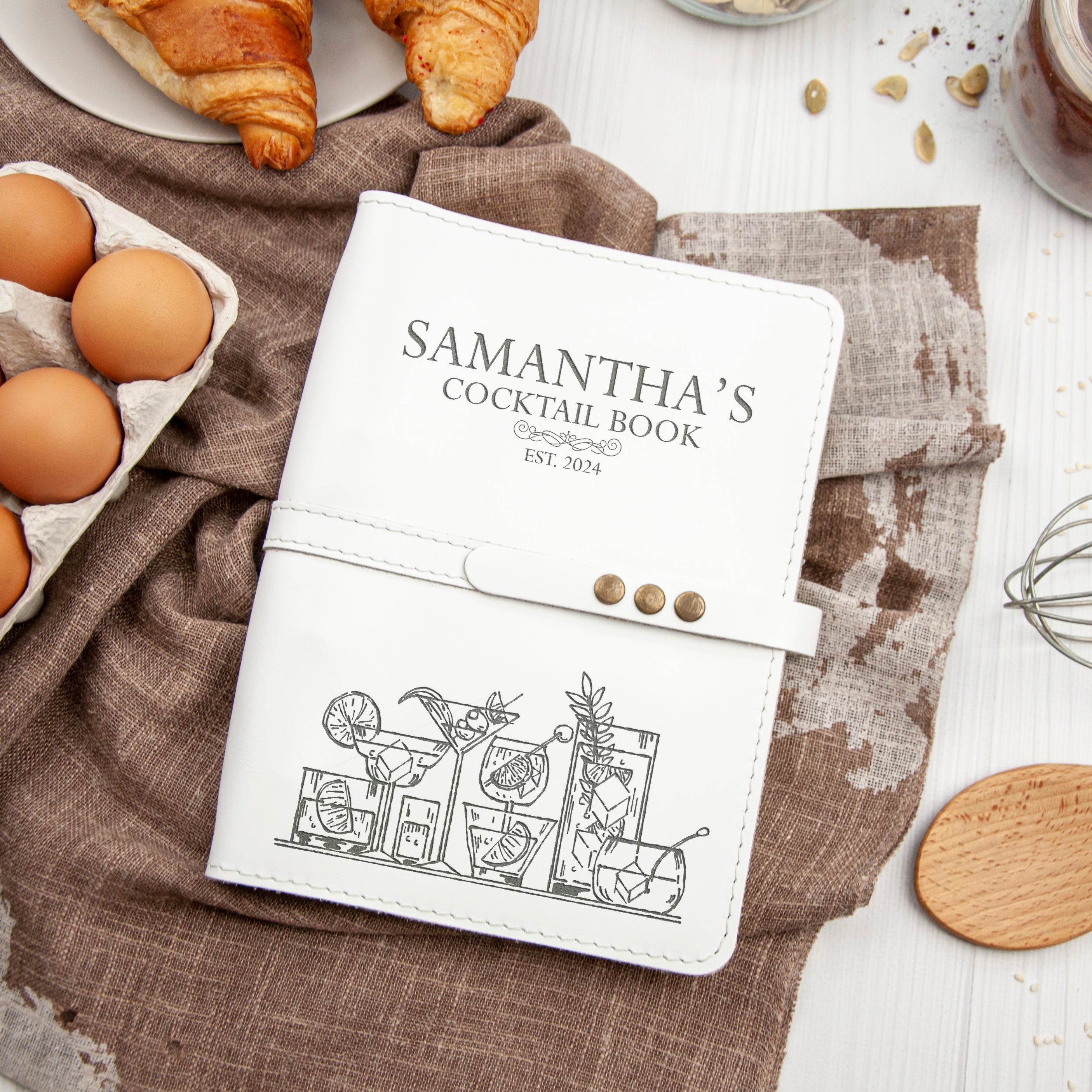 A white cocktail book with a design of cocktail glasses and a cocktail on the cover, placed on a table with various food items such as eggs, a croissant, and a wooden spoon.