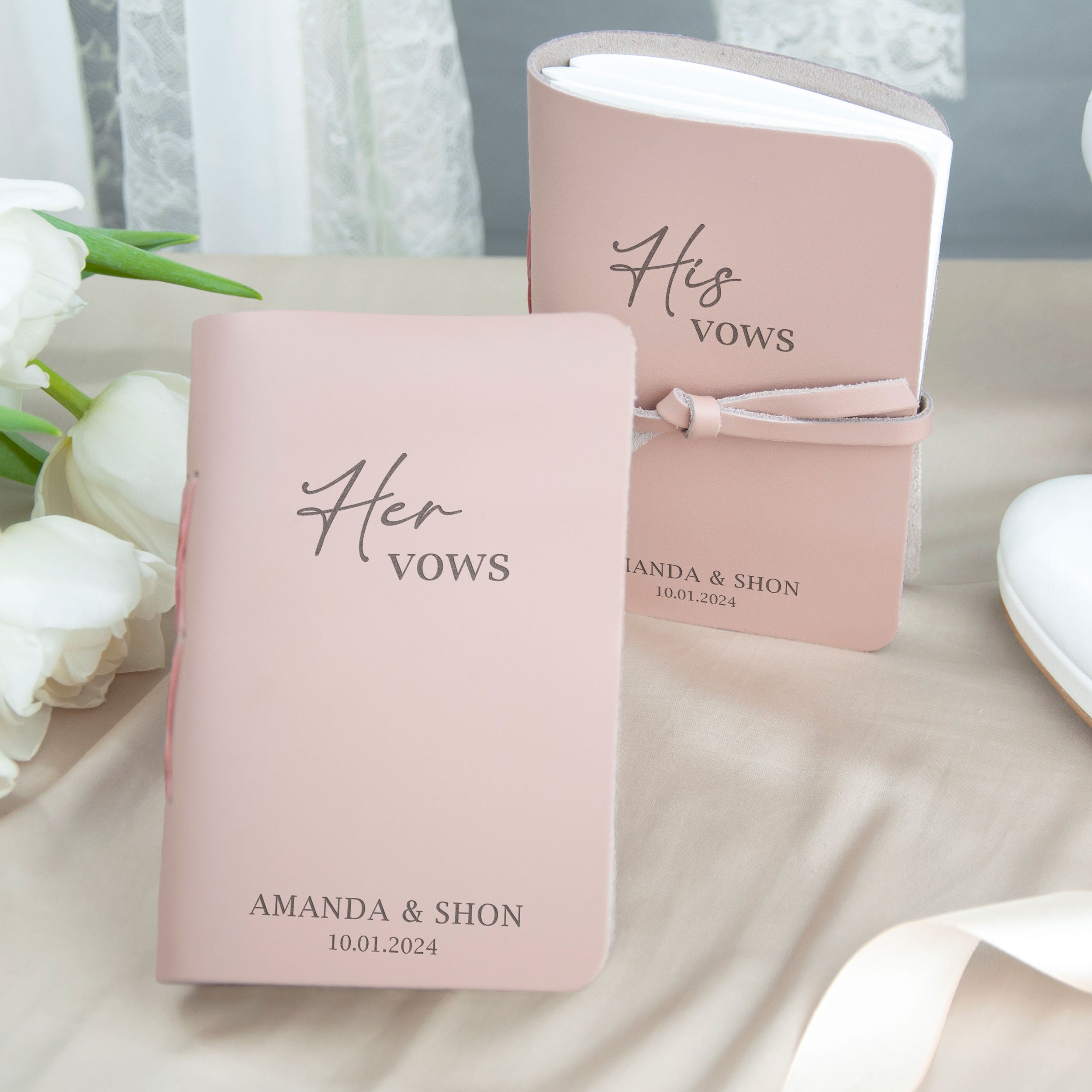 two pink wedding vows books with the text "Her Vows" printed on them, placed on a table with white roses in the background.