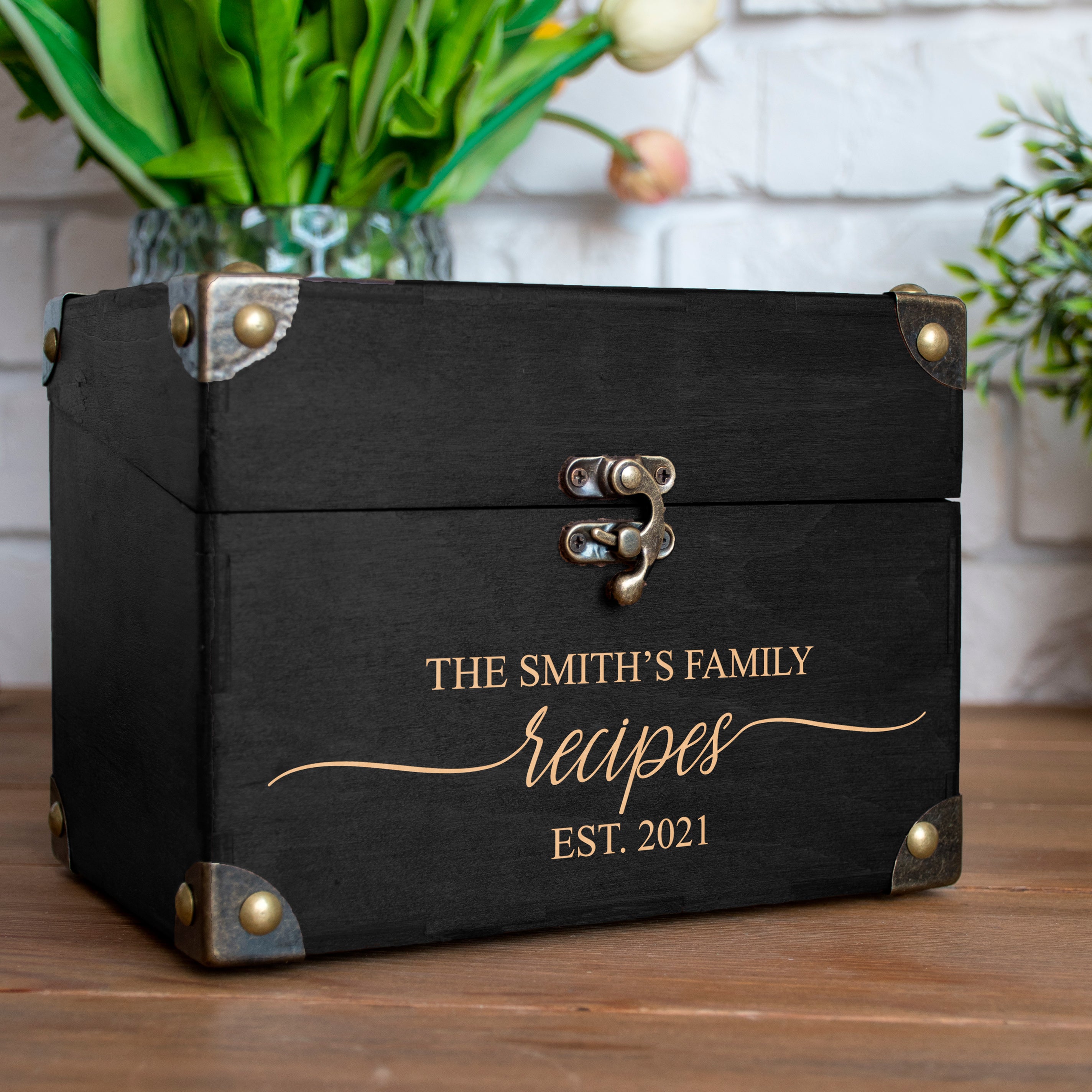 A black wooden recipe box with brass hardware and a gold cursive inscription, placed on a wooden surface with a vase of green leaves in the background.