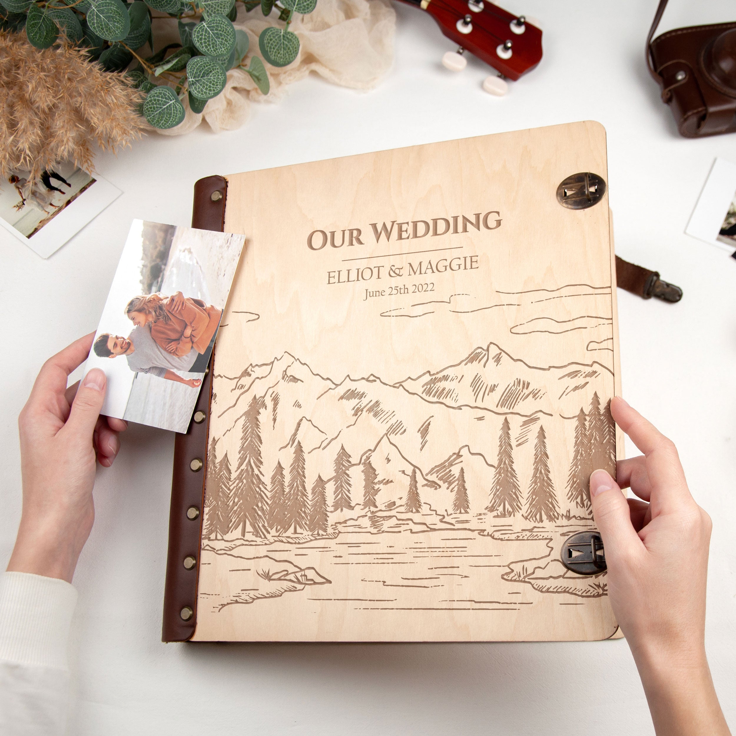 A person is holding a wooden wedding album with a mountain landscape engraved on the cover, and there is a photograph attached to the album.