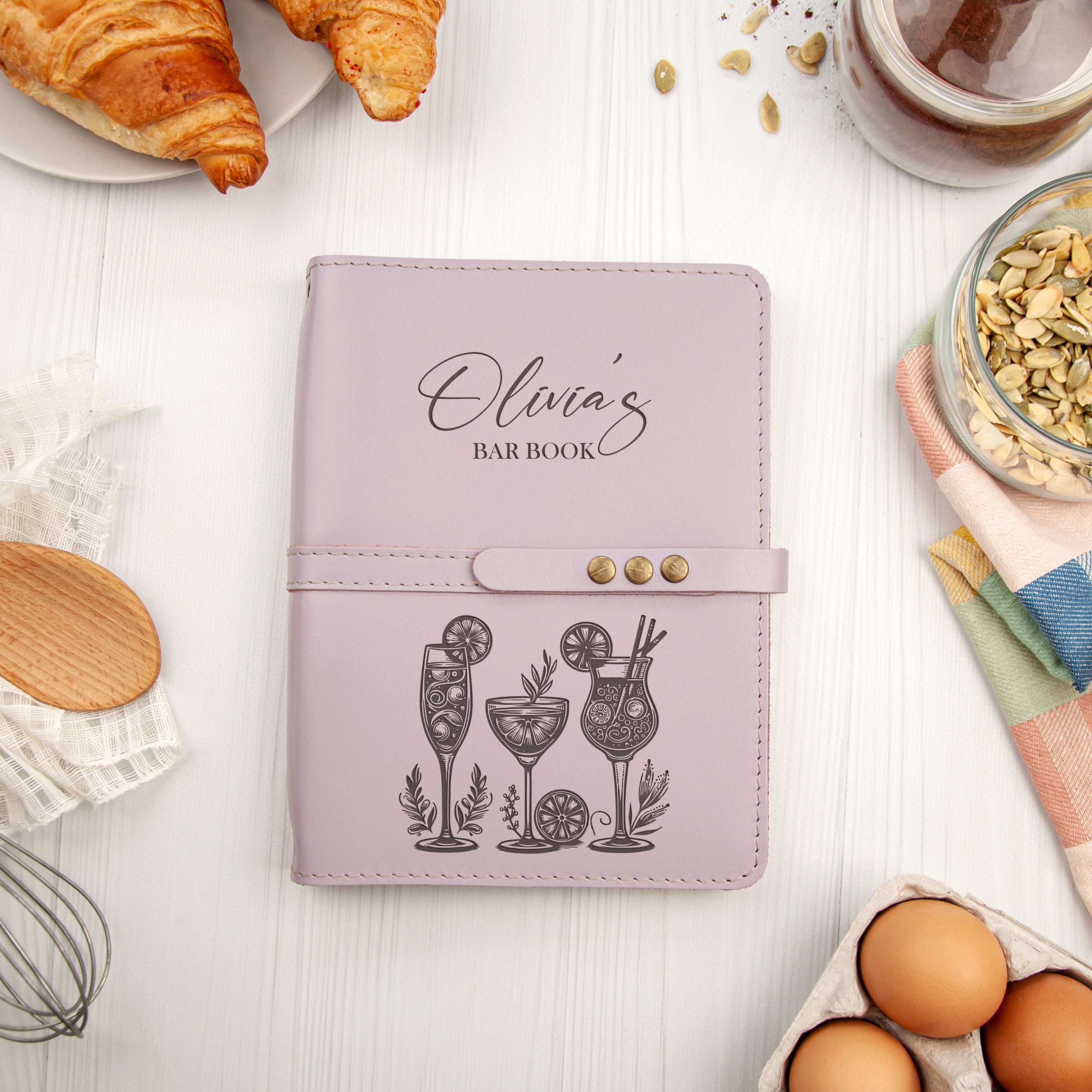 a pink leather-bound notebook with a personalized design, surrounded by various food items and kitchen utensils on a white surface.