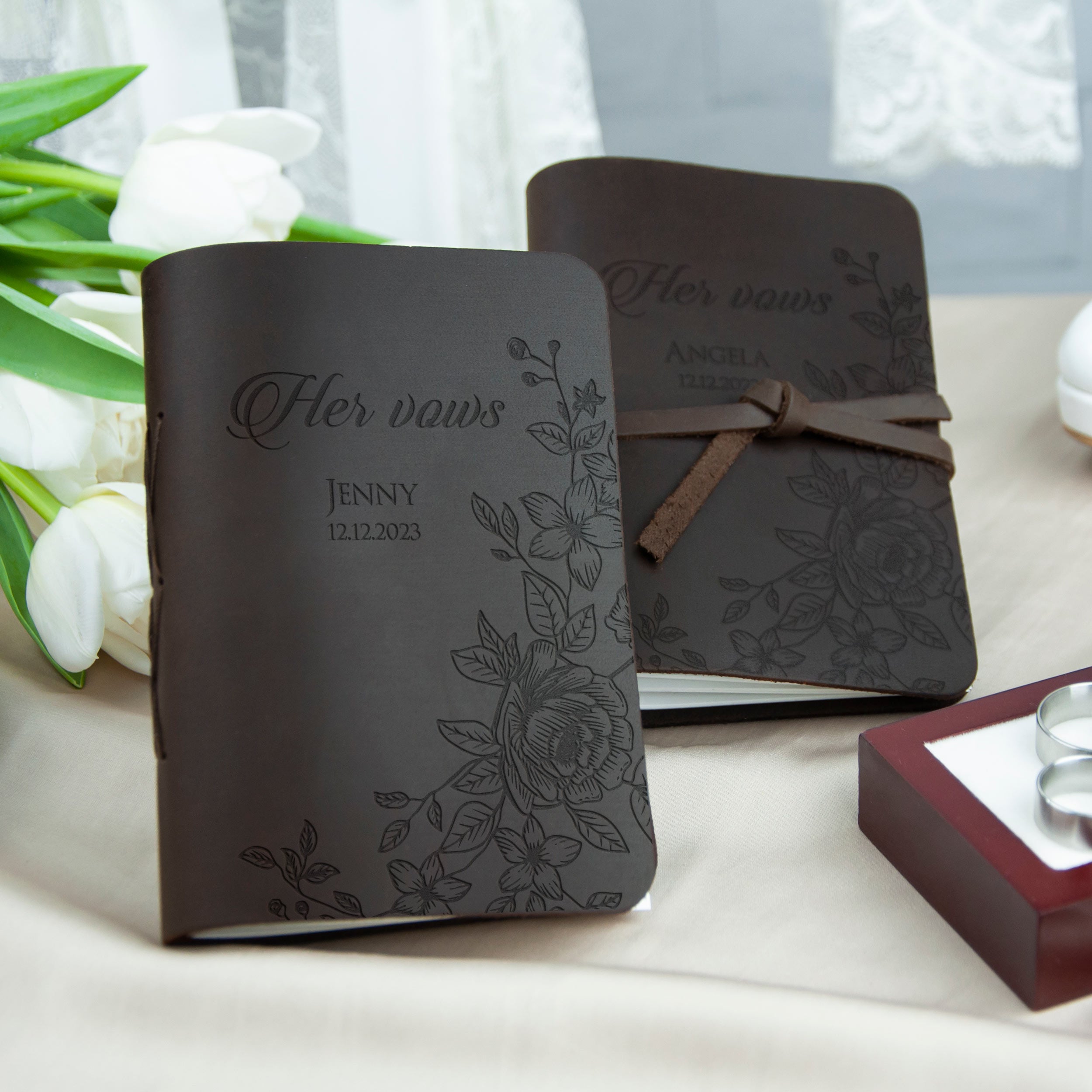 two black leather-bound books with floral designs, placed on a table with white flowers in the background.