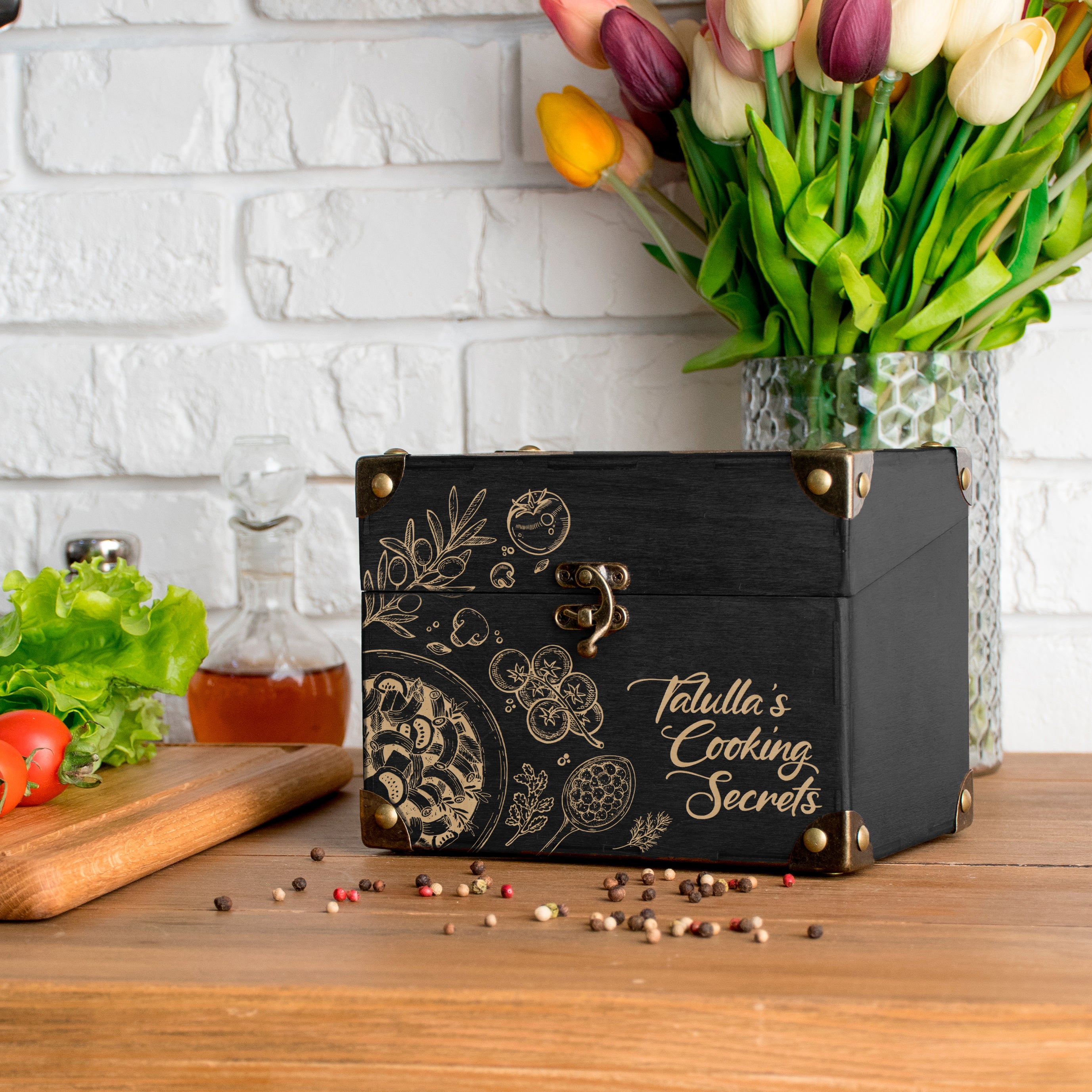 A black wooden box with gold designs and text, placed on a wooden table next to a vase of tulips, with a cutting board and spices nearby.