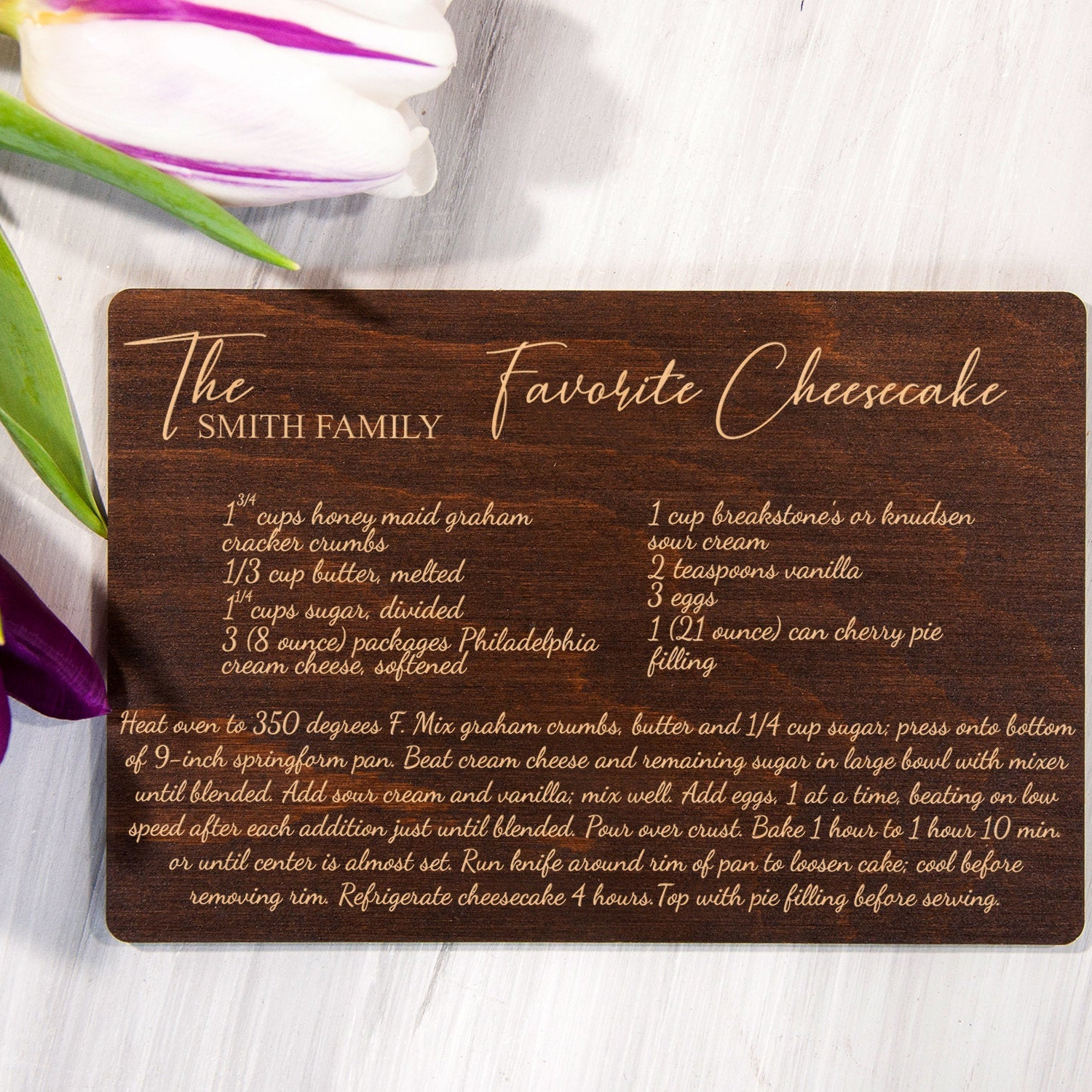 A wooden coaster with a recipe for "The Smith Family's Favorite Cheesecake" on it, along with some flowers and a knife.