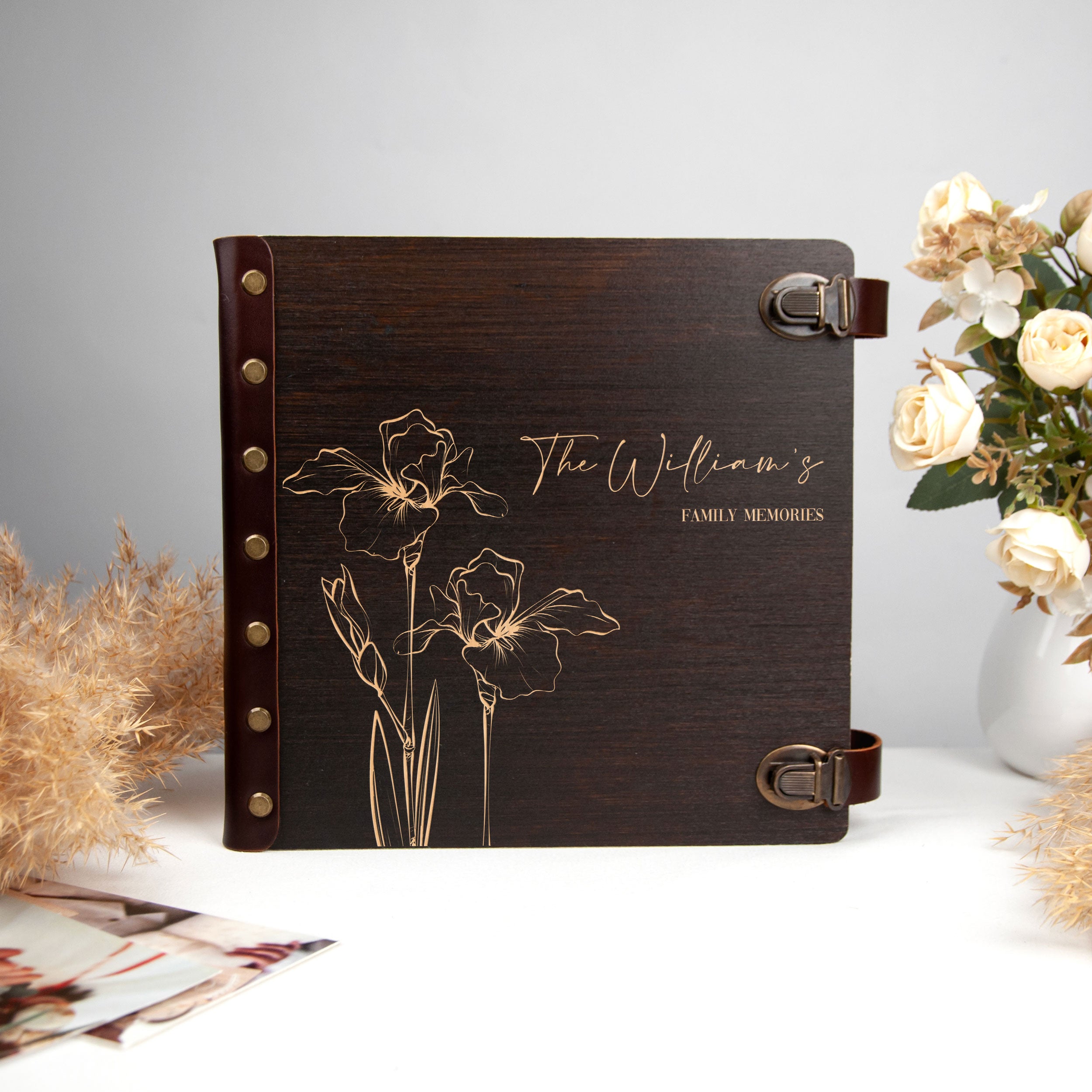 A dark brown photo album with gold floral designs, placed on a white surface next to a vase of white flowers.