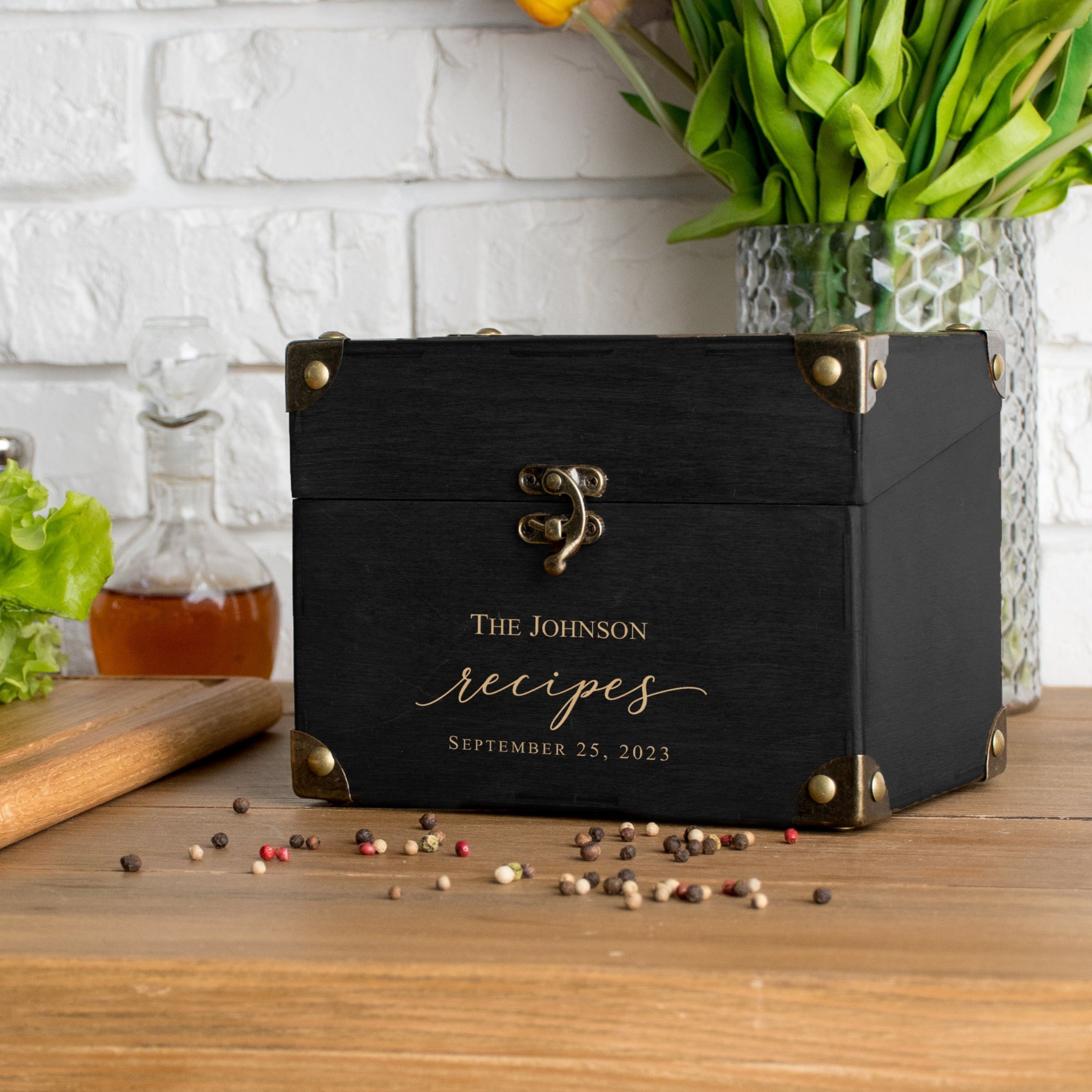 a black wooden box with gold accents, labeled "The Johnson Recipes September 25, 2023", placed on a wooden table next to a vase of colorful tulips.