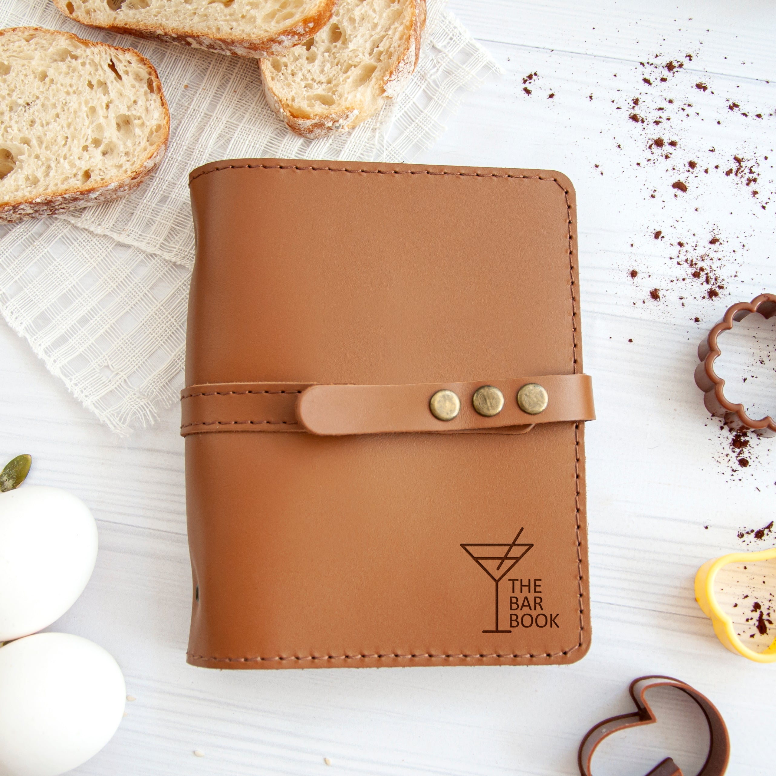 A brown leather-bound notebook with a cocktail glass design sits on a white surface, surrounded by various food items and kitchen tools.