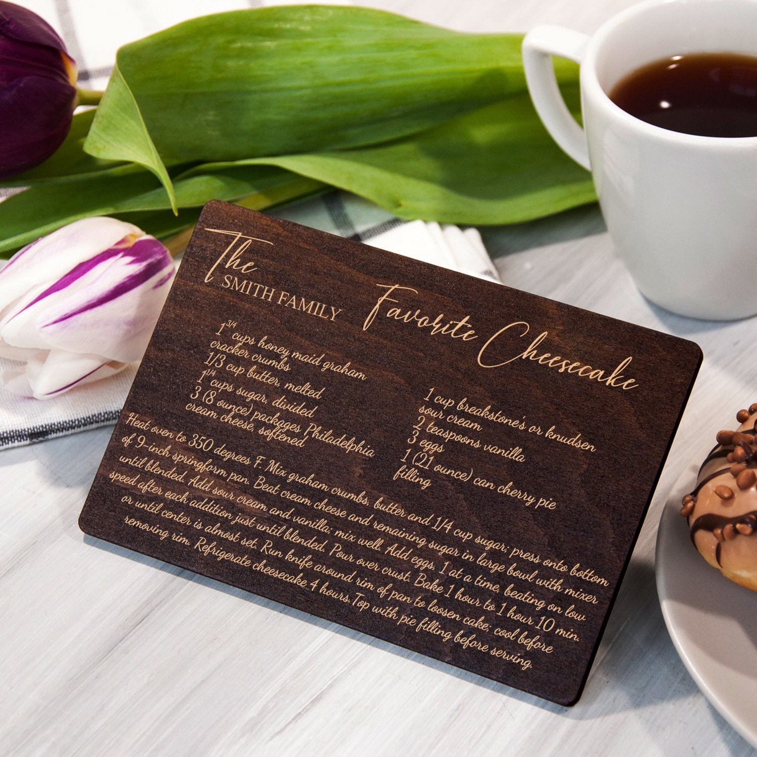 A wooden recipe card titled "The Smith Family Favorite Cheesecake" is placed on a table next to a cup of coffee and a chocolate donut.
