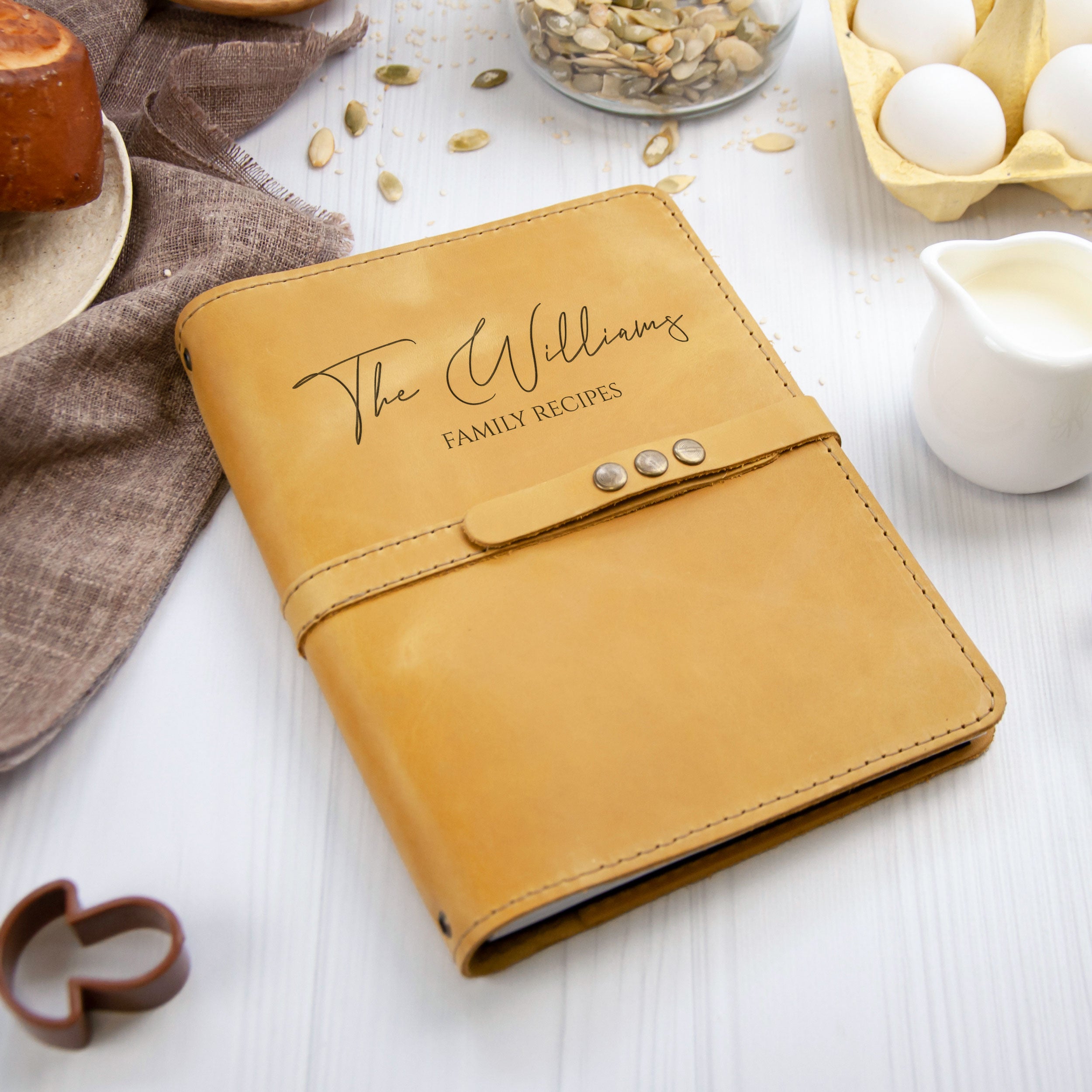 A tan leather-bound recipe book titled "The Williams Family Recipes" sits on a white surface, surrounded by various kitchen items such as a carton of eggs, a glass jar, and a white pitcher.