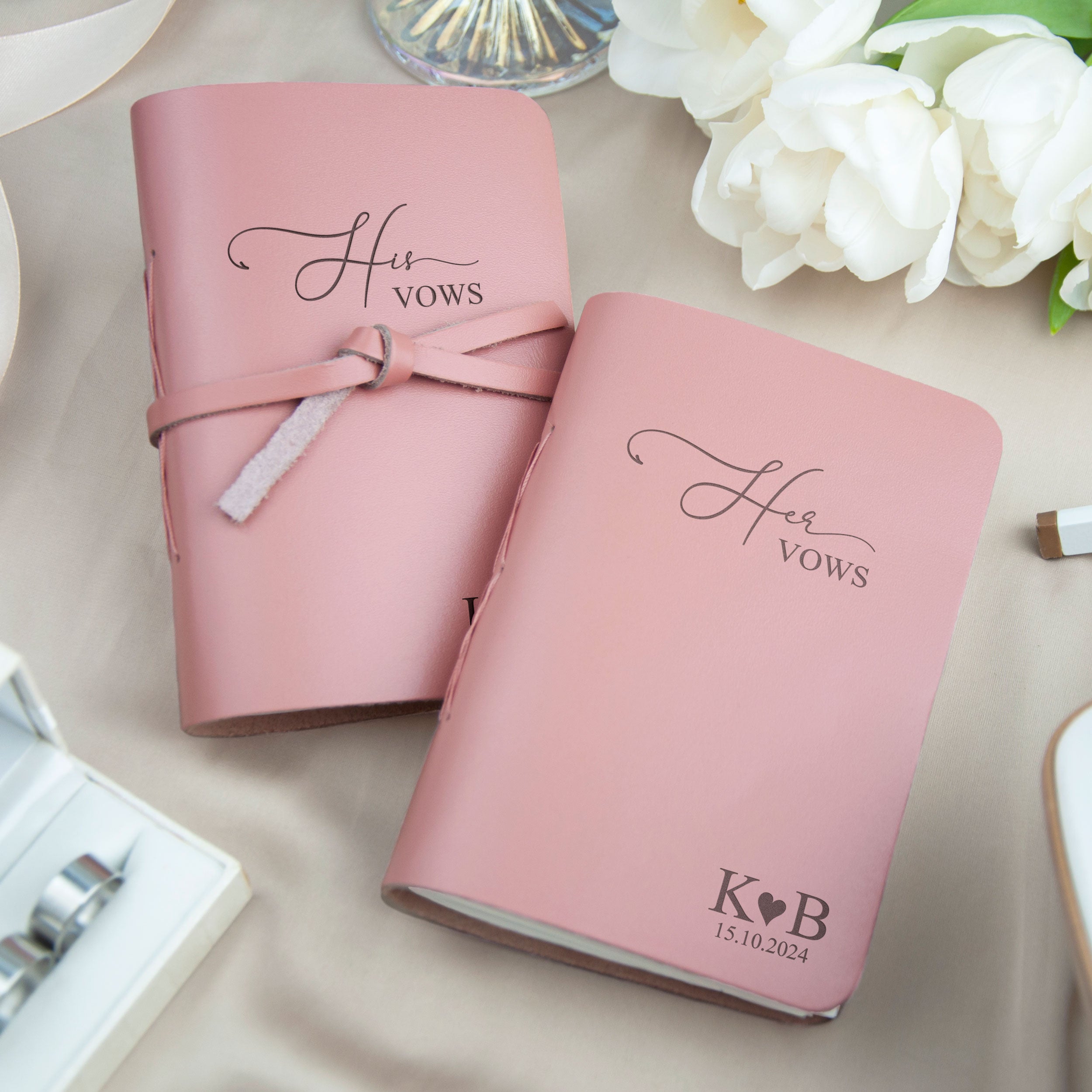 Two pink leather-bound notebooks with the text "Hrs Vows" printed on them, along with a small floral arrangement and a ring box in the background.