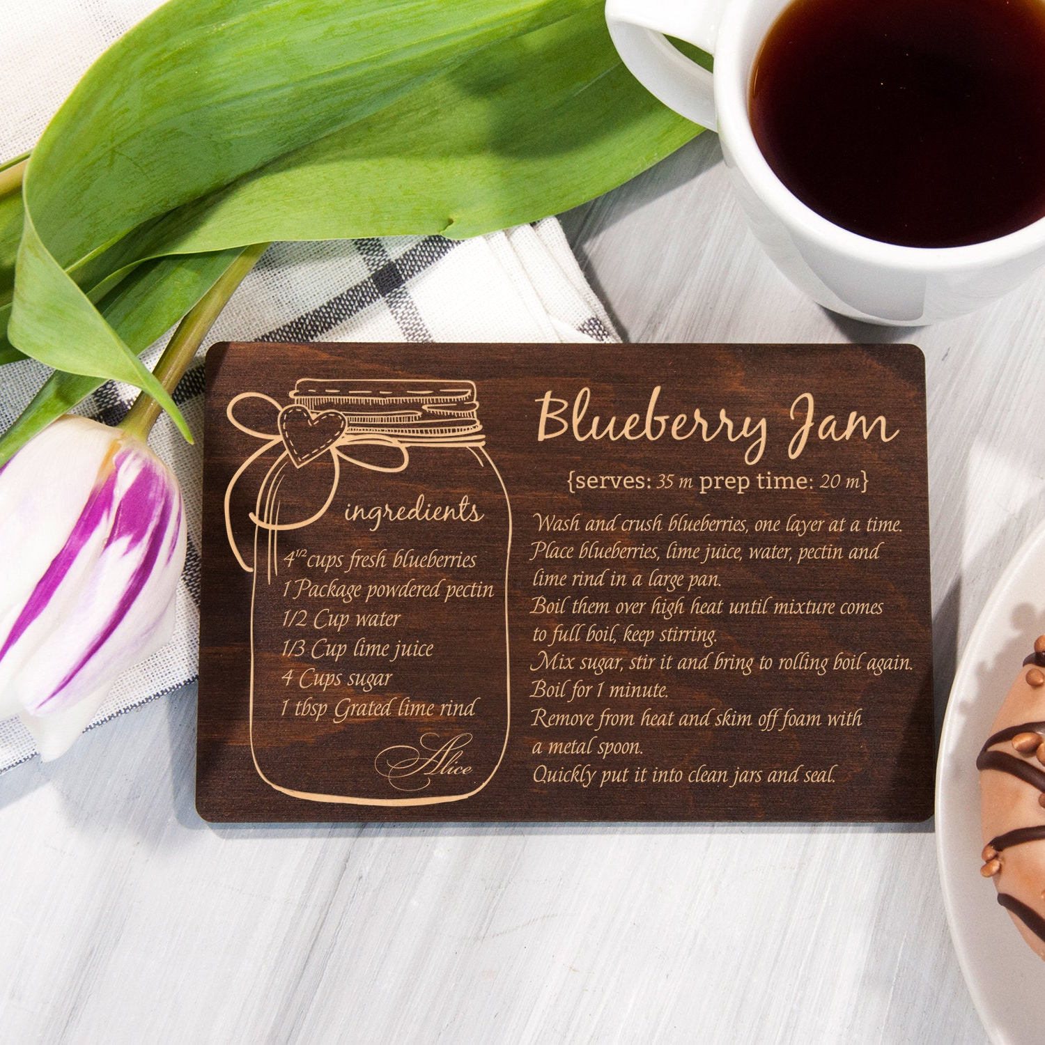 A wooden coaster with a recipe for Blueberry Jam is placed on a table next to a cup of coffee and a pastry.