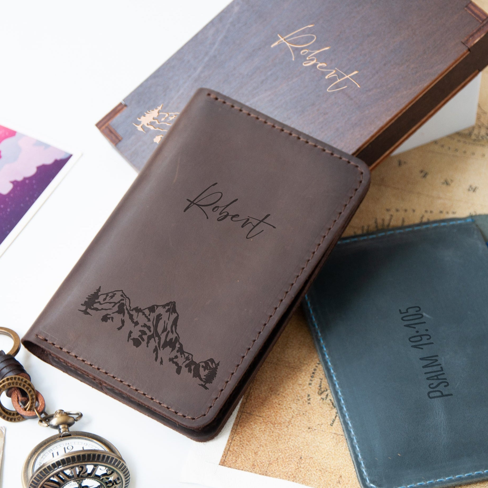 a leather wallet with a mountain landscape design, a small blue wallet, and a pocket watch.