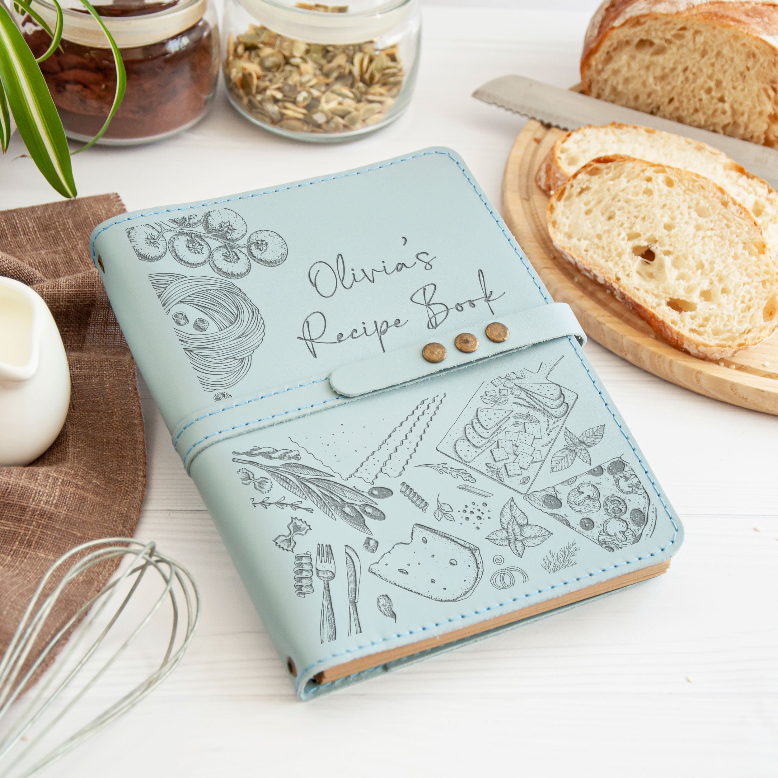 A light blue recipe book with illustrations of food items and a personalized name on the cover, placed on a wooden surface alongside a loaf of bread, a knife, and some jars of food.