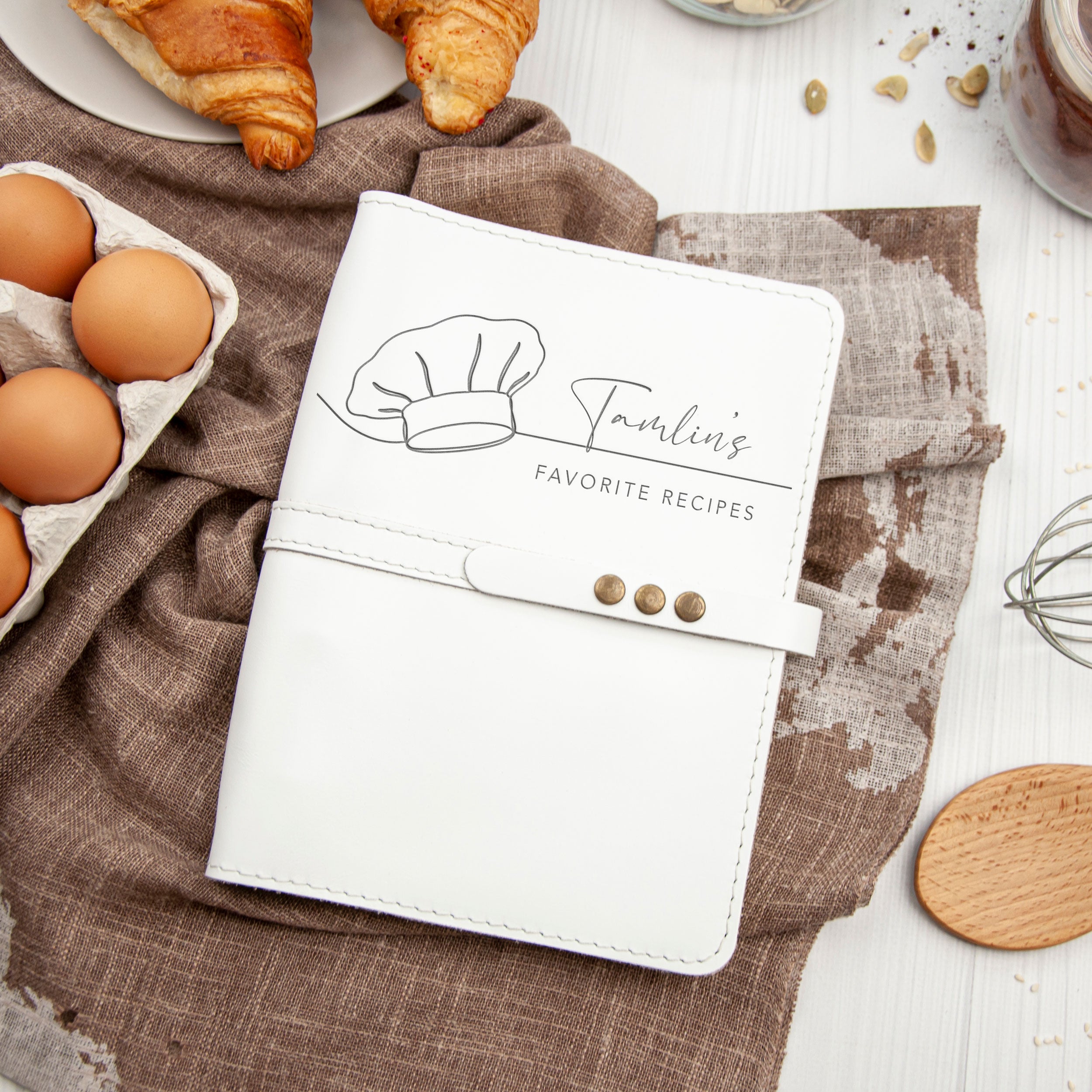 A white cookbook with a chef's hat and the text "Tulini's Favorite Recipes" is placed on a table with various food items and kitchen utensils.