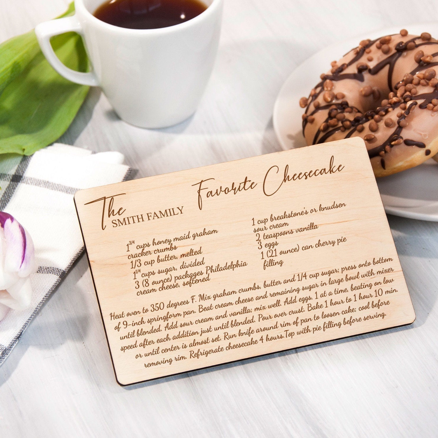 A wooden recipe card titled "The Smith Family Favorite Cheesecake" is placed on a table next to a plate of chocolate-covered donuts and a cup of coffee.