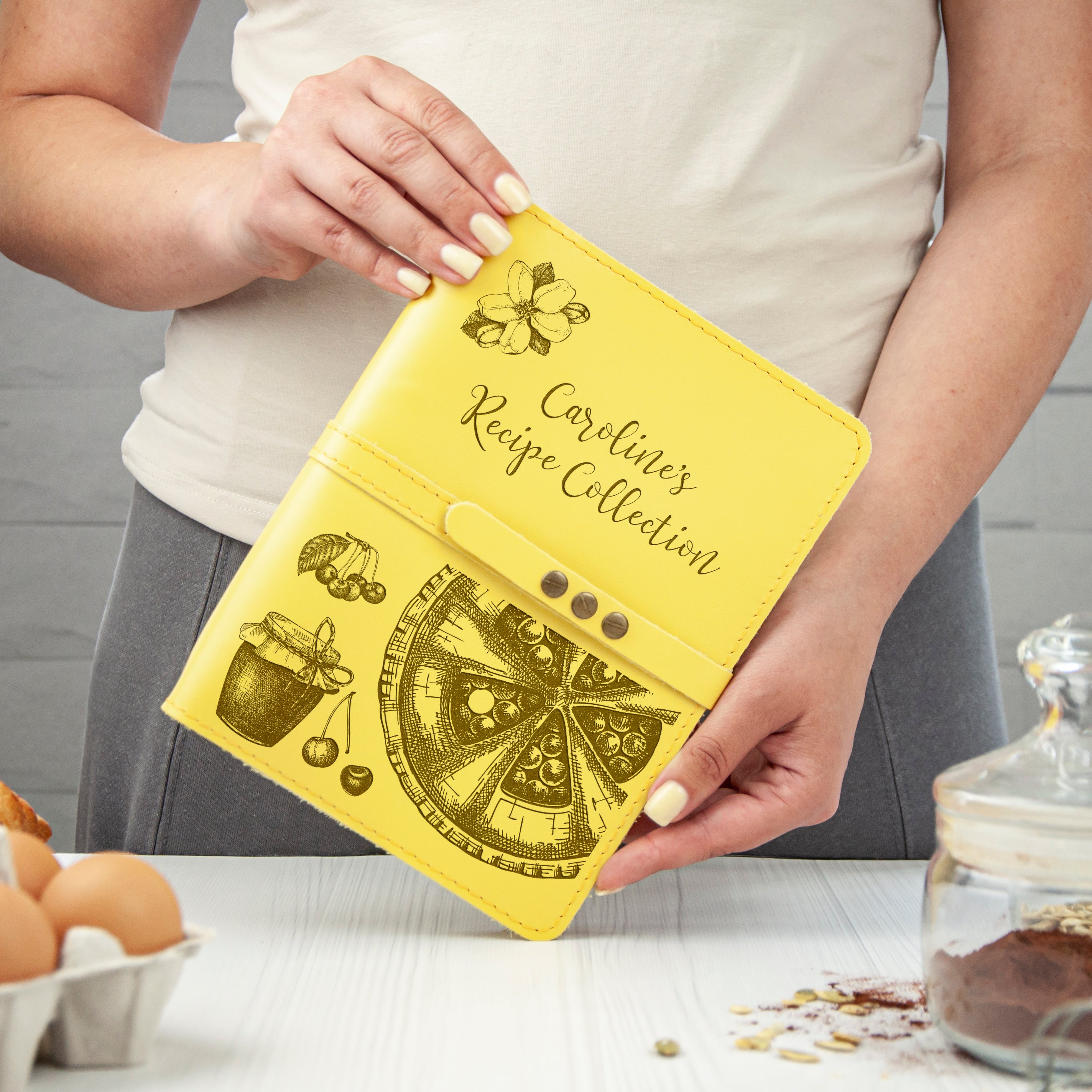 A person is holding a yellow recipe book with illustrations of food items, such as a pizza and a jar of jam, and the title "Cardliner's Recipe Collection" printed on it.