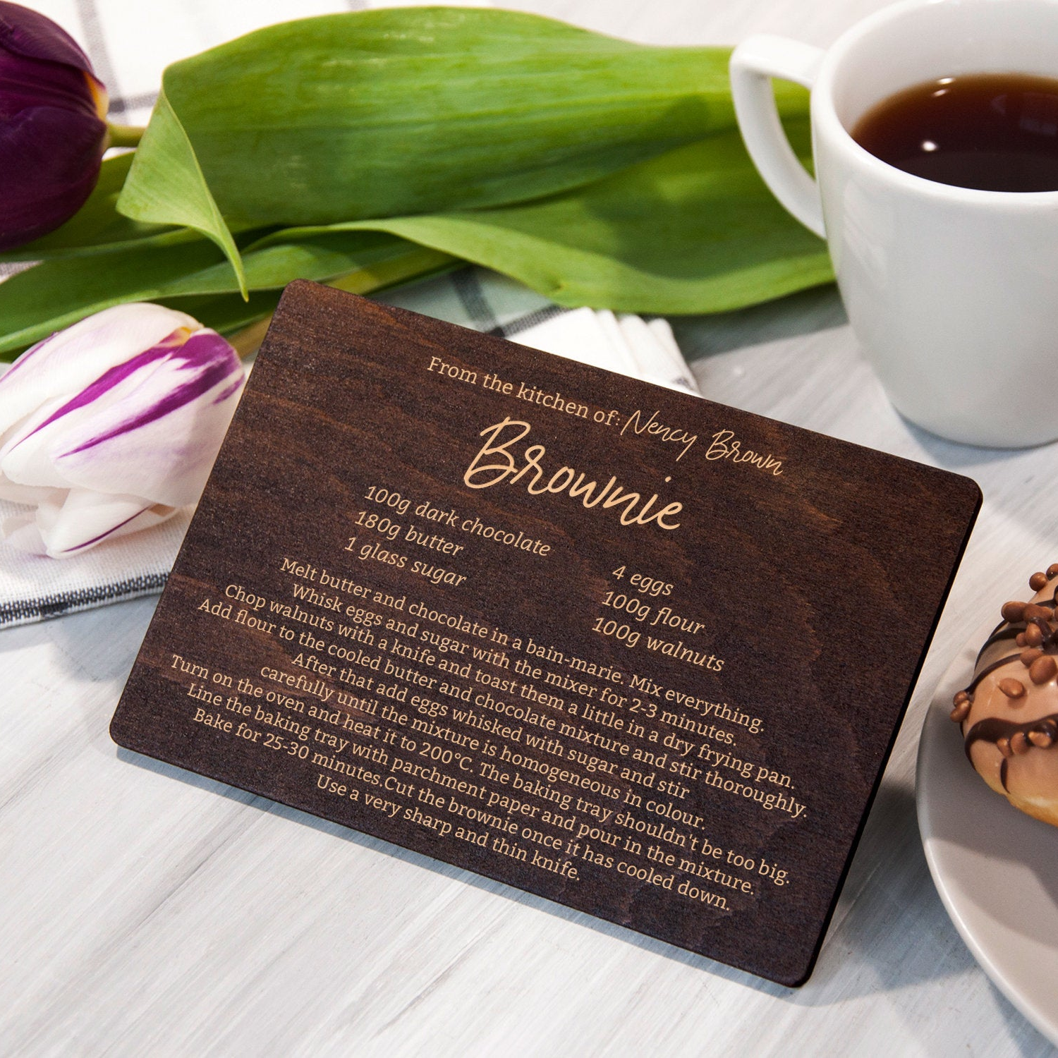 A wooden recipe card for "Brownie" is placed on a table next to a cup of coffee and a plate with a donut.