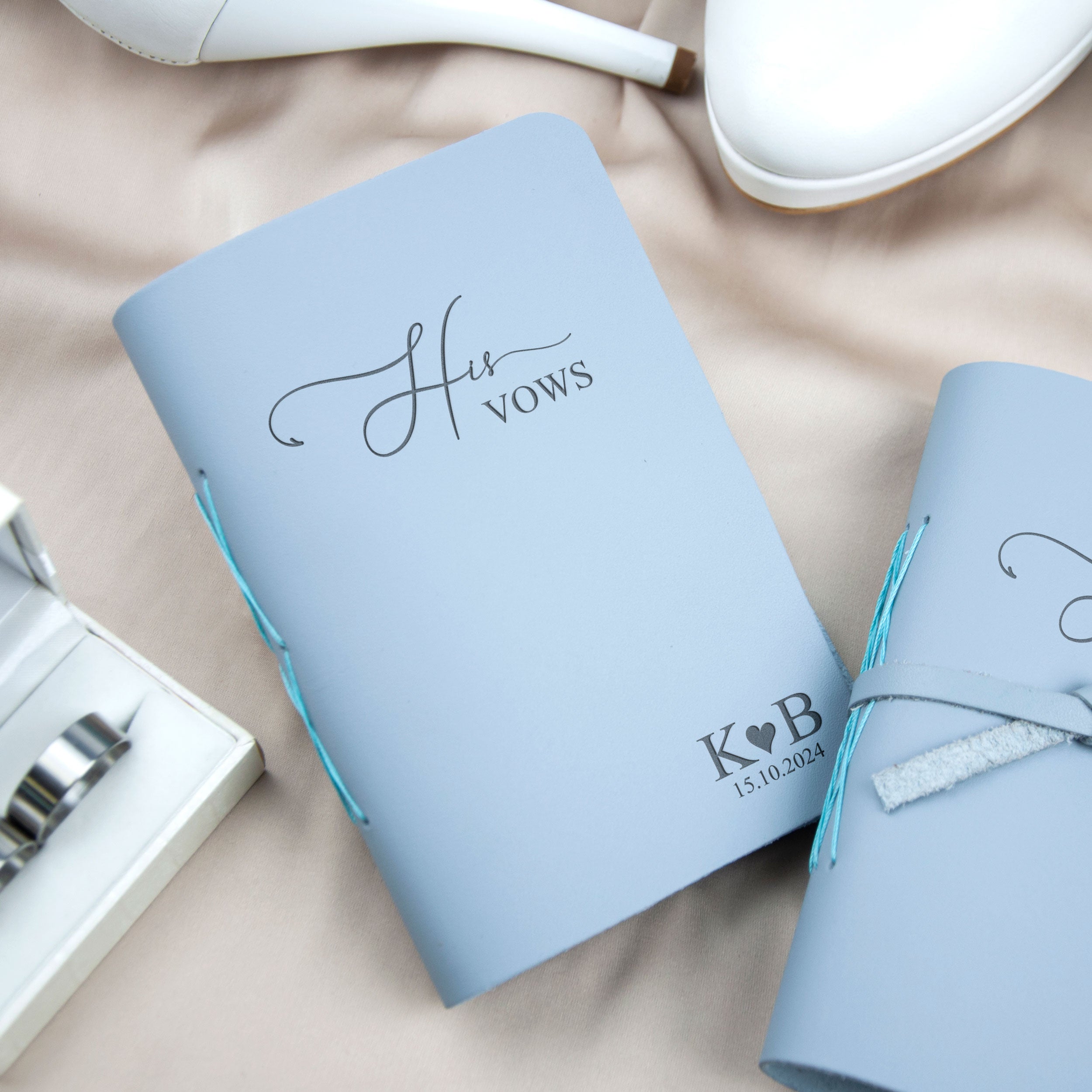 A light blue notebook with the text "His Vows" printed on it, along with a small white shoe and a silver ring, all placed on a beige surface.