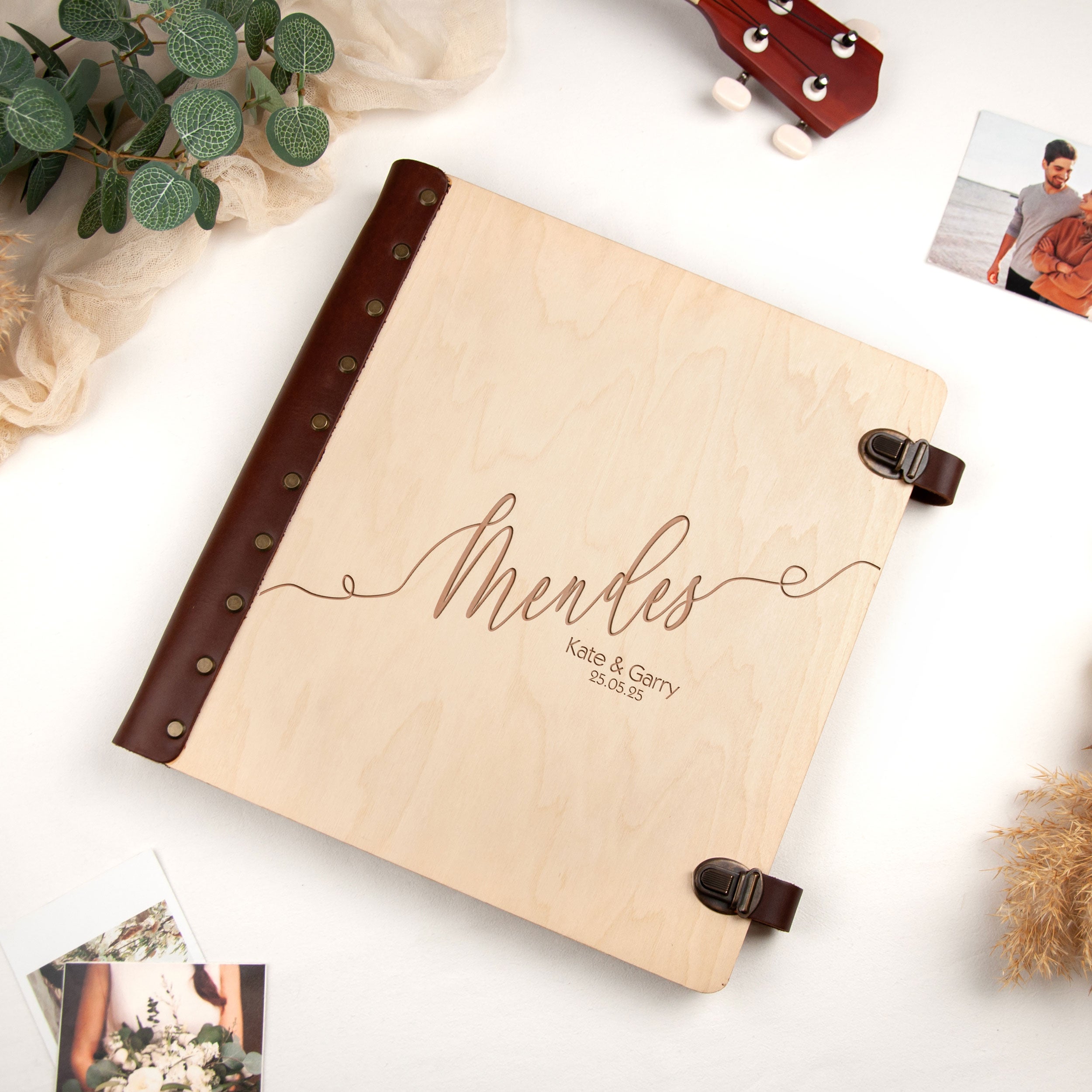 a wooden album with a leather cover, displaying the name 'Mendes' and the names 'Kate & Gary' on its cover. The album is placed on a white surface, surrounded by various items such as a guitar, a plant, and some photographs.