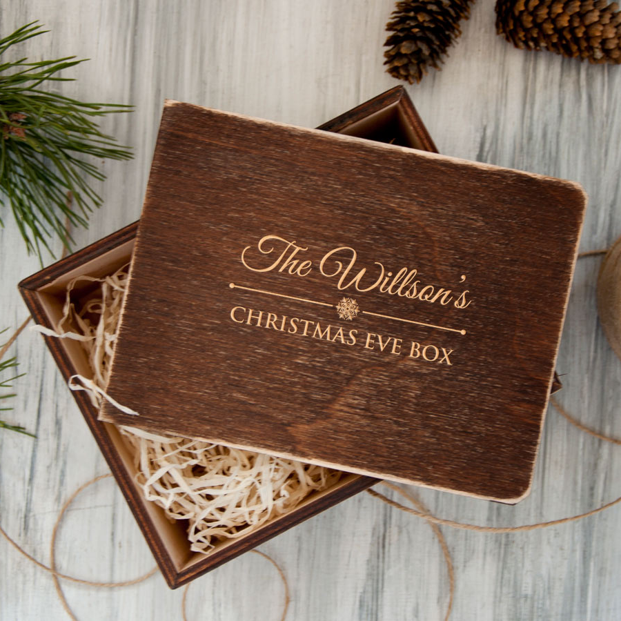 A wooden box with the text "The Wilson's Christmas Eve Box" printed on it, placed on a wooden surface with pine cones and greenery in the background.