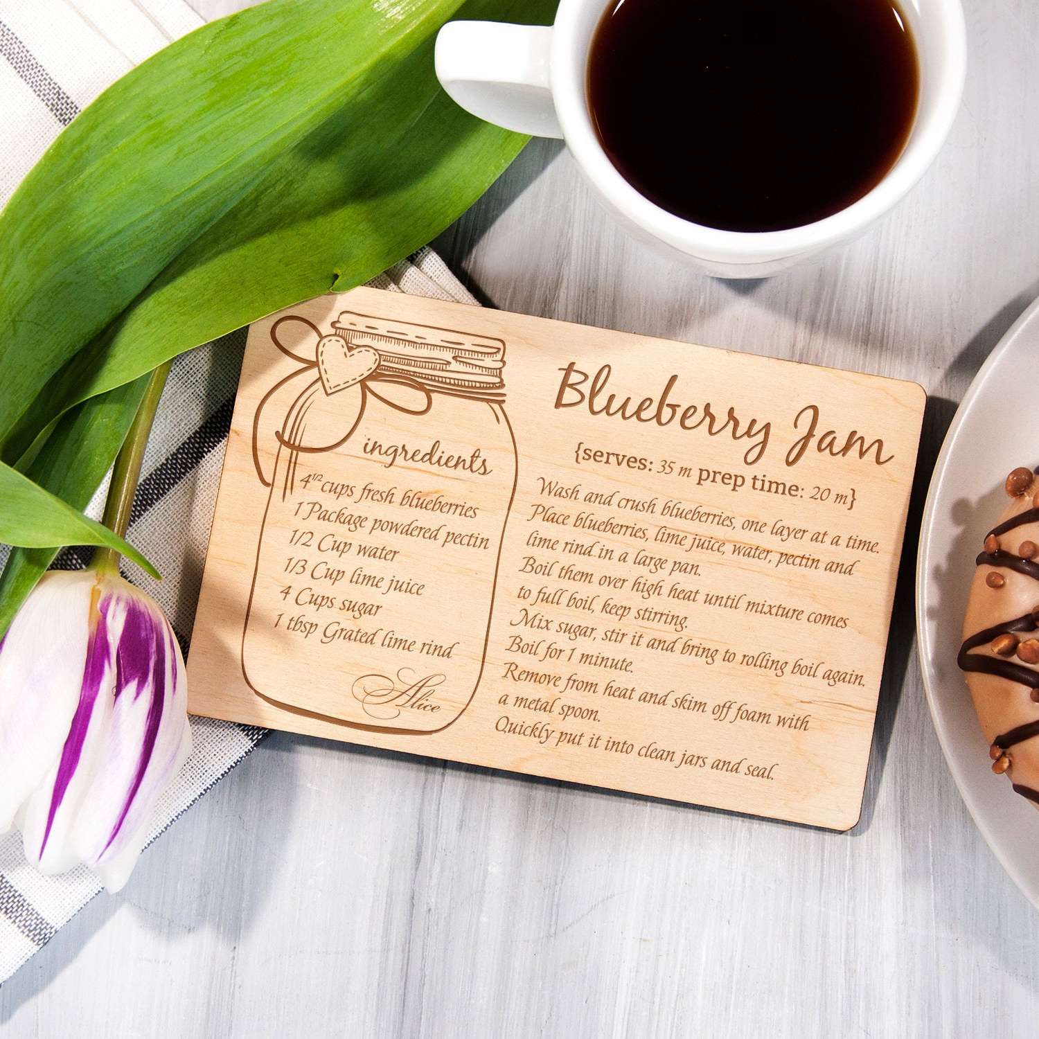 A wooden recipe card with a blueberry jam recipe is placed on a table next to a cup of coffee and a plate of donuts.