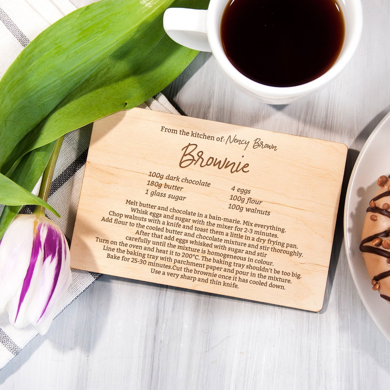 A wooden coaster with a recipe for "Brownie" from the kitchen of Nancy Brown, accompanied by a cup of coffee and a plate of what appears to be a chocolate dessert.