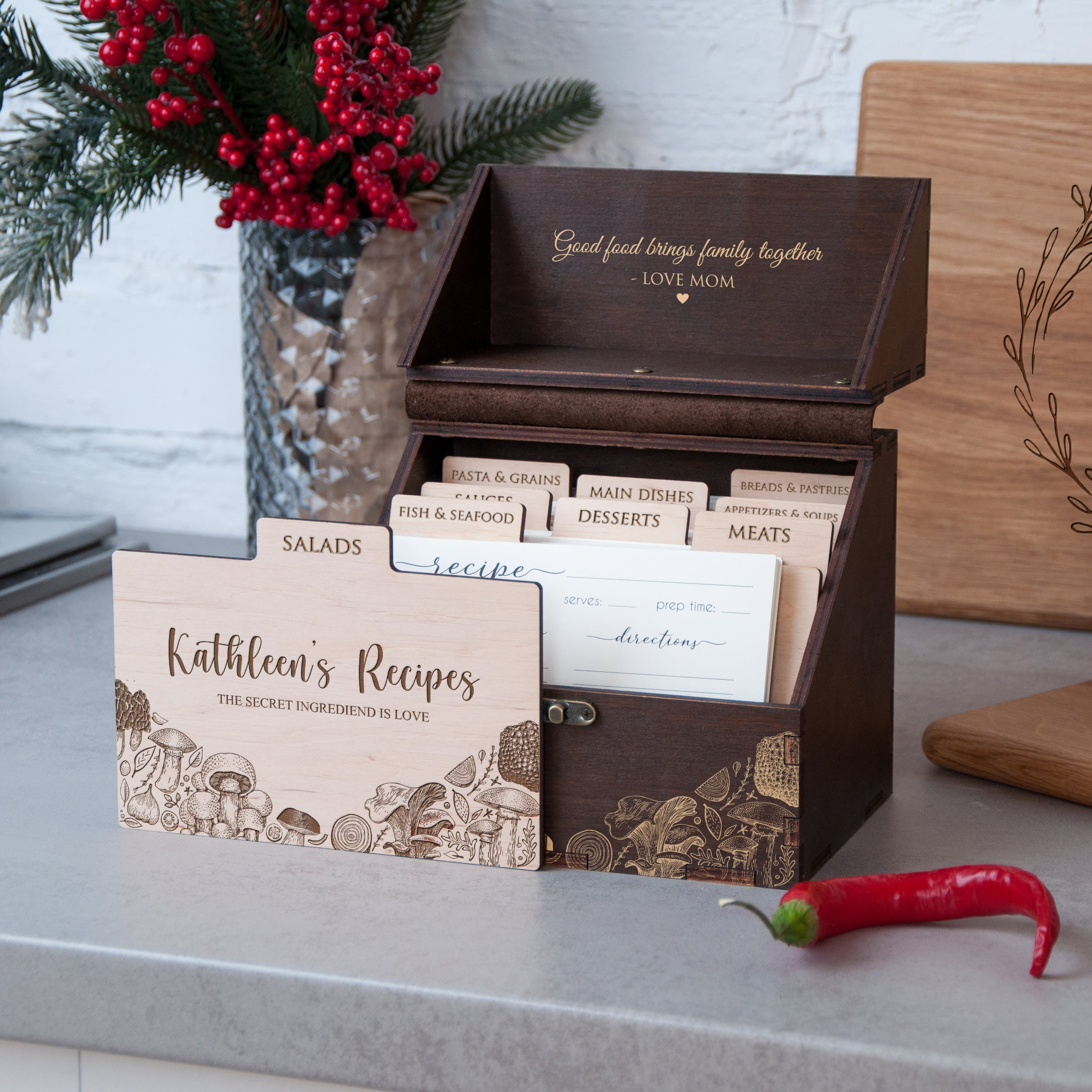 A wooden recipe box with a lid, displaying a list of recipes, sits on a countertop next to a vase of red berries and a wooden cutting board.
