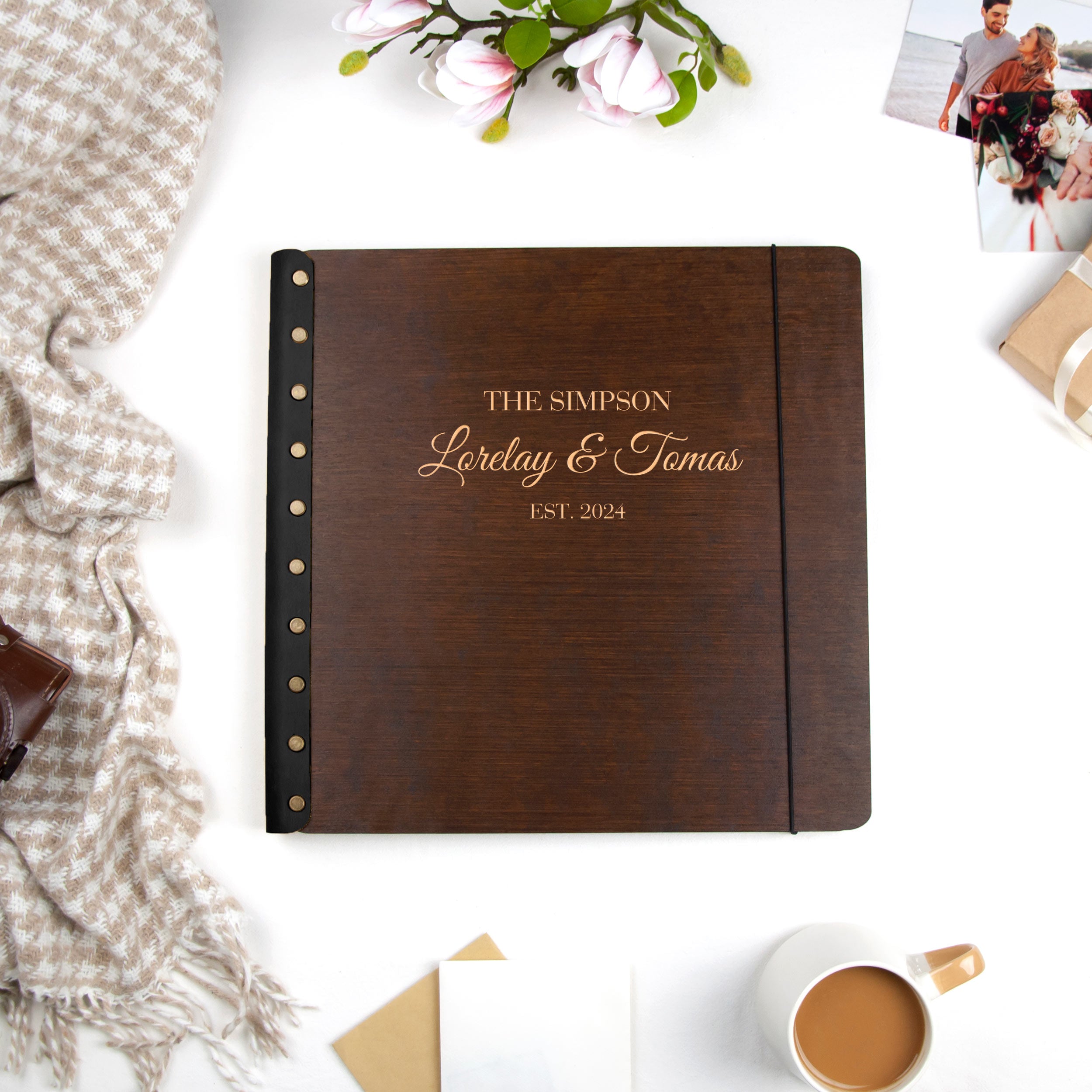a wooden photo album with the text "THE SIMPSON Lorelay & Tomas EST. 2014" printed on it, placed on a white surface alongside other items such as a mug, a flower, and some photographs.