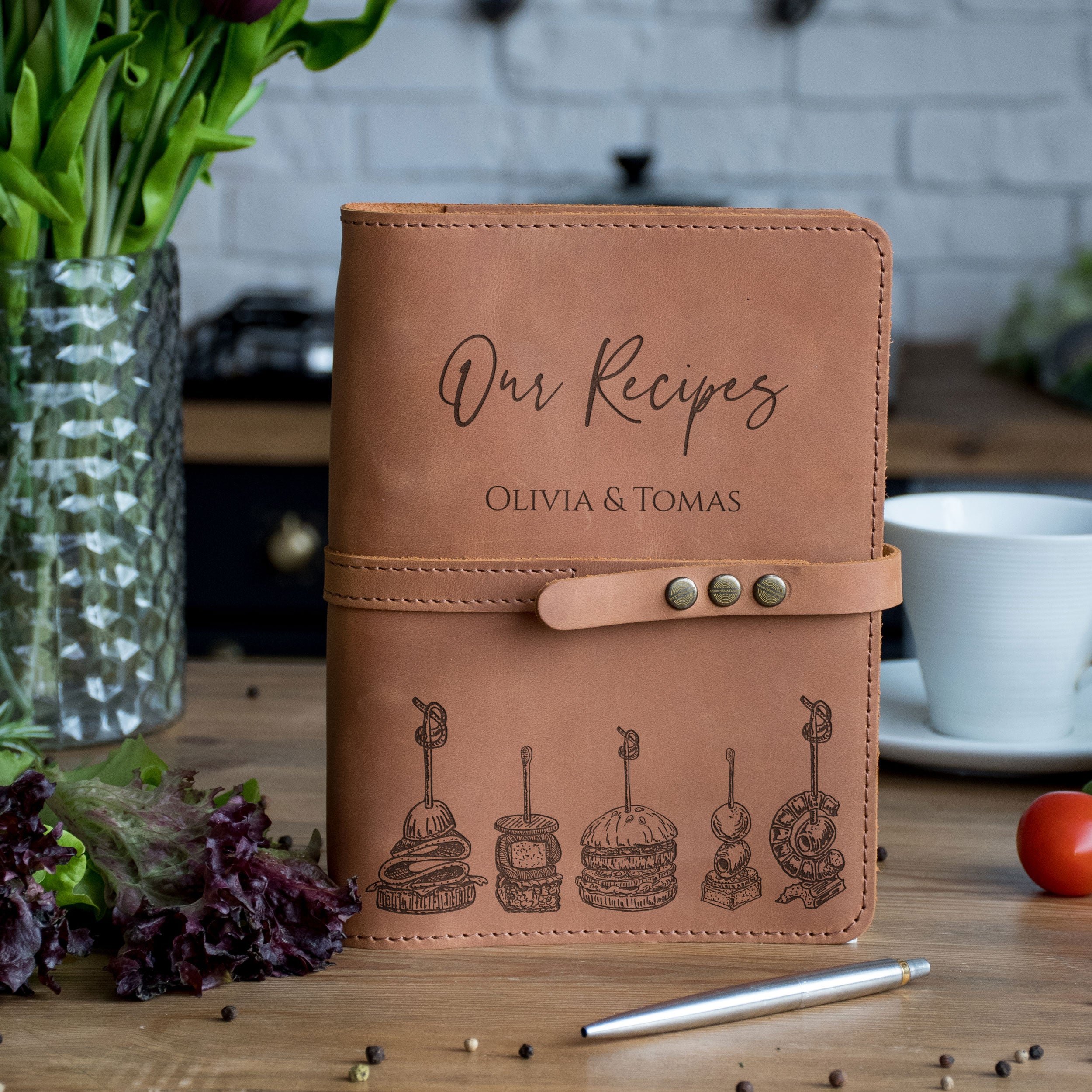 A leather-bound recipe book titled "Our Recipes" by Olivia & Tomas, featuring illustrations of various food items, sits on a wooden table surrounded by a vase of flowers, a cup, and a pen.