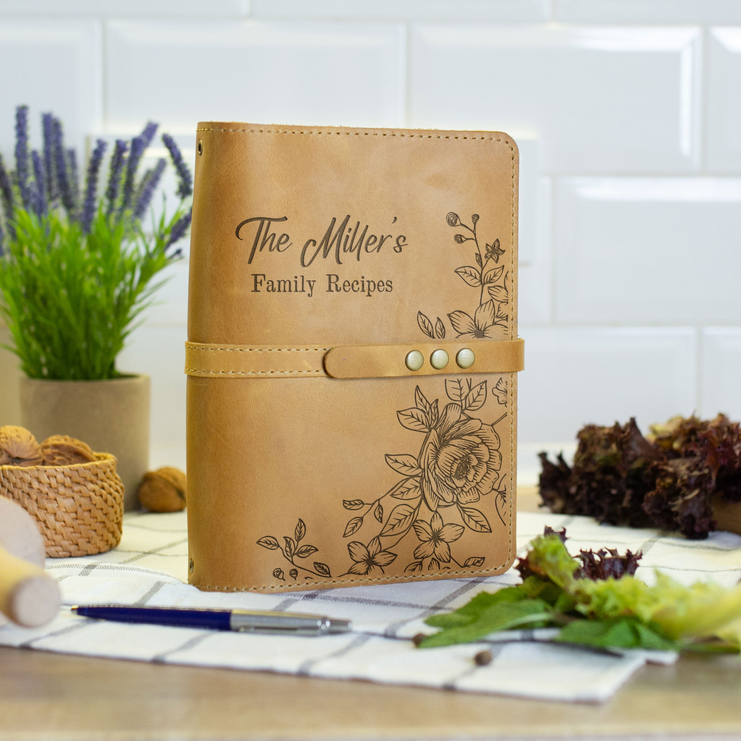 A leather-bound notebook with the title "The Miller's Family Recipes" printed on it, placed on a table with a pen and a plant in the background.