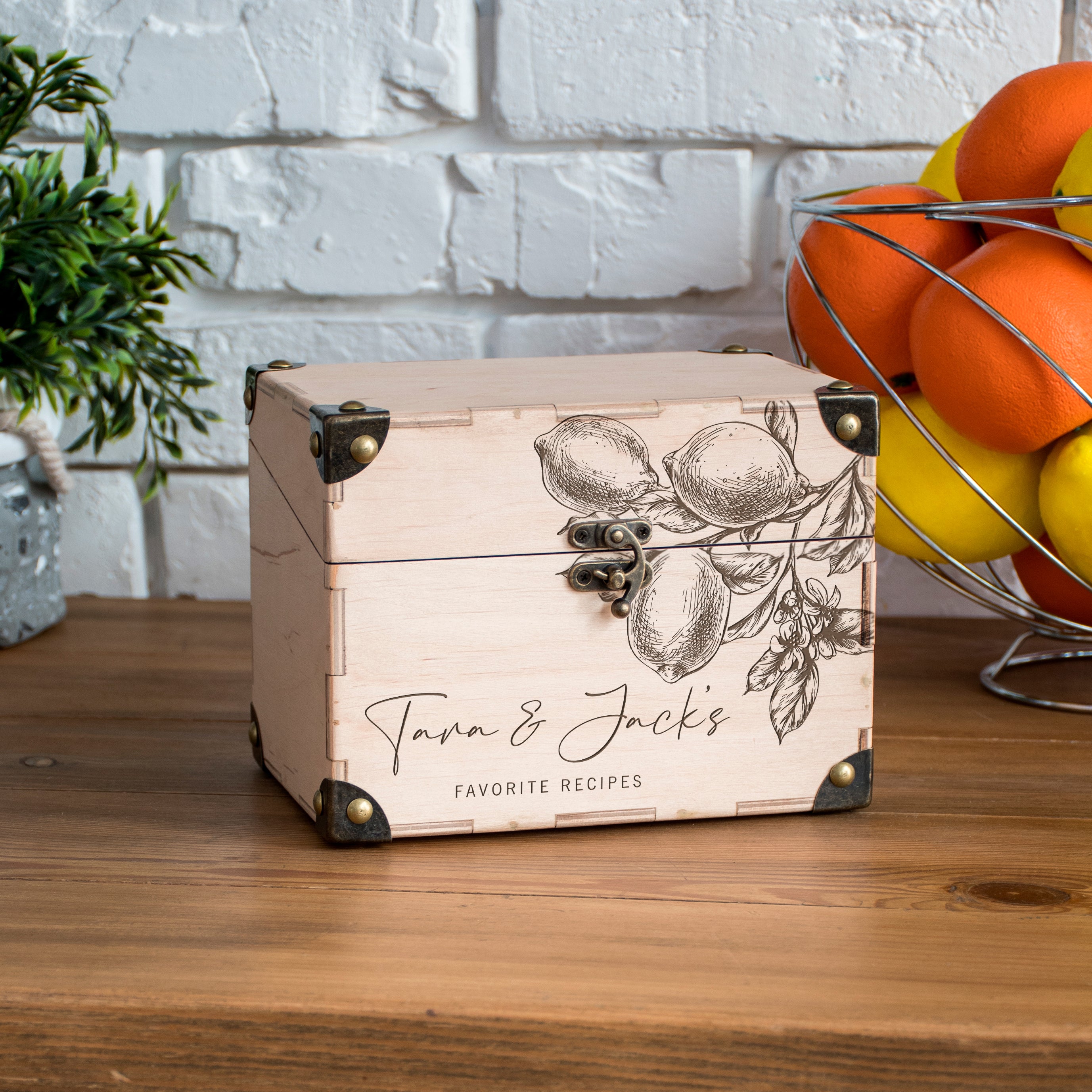 A wooden box with a decorative design featuring lemons and oranges, placed on a wooden surface next to a wire fruit basket filled with oranges and lemons.