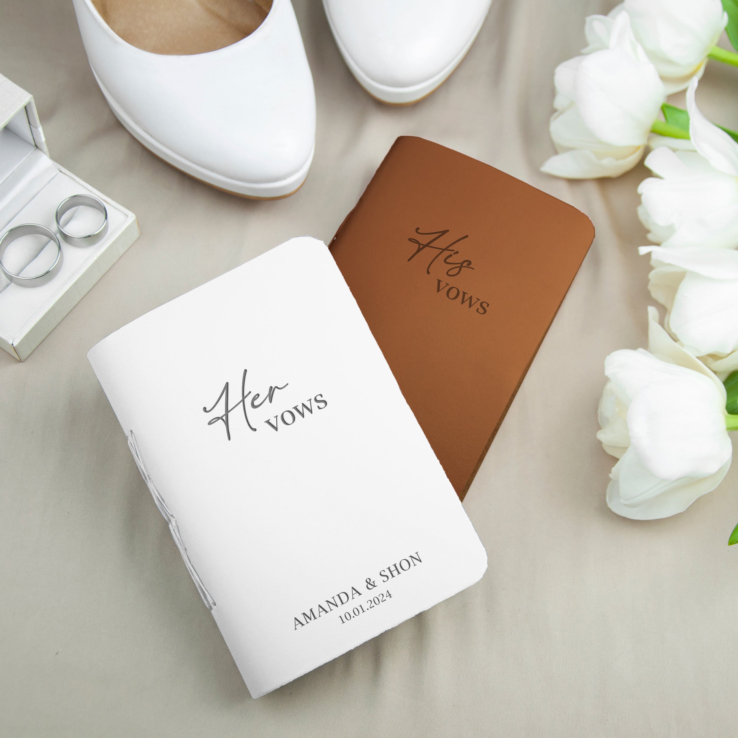 a pair of white and brown wedding vows books, a pair of white wedding rings, and a pair of white shoes, all arranged on a beige background.