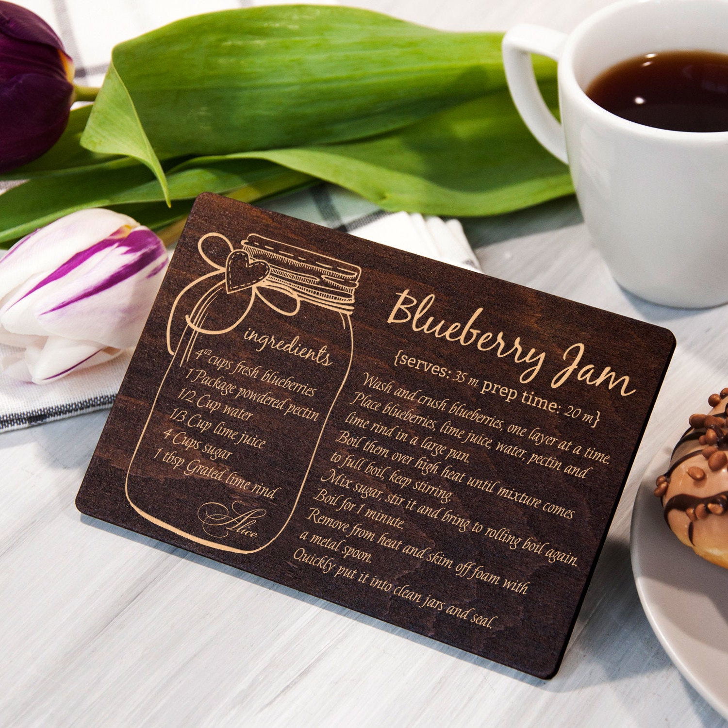 A wooden recipe card with a blueberry jam recipe is placed on a table next to a cup of coffee and a pastry.