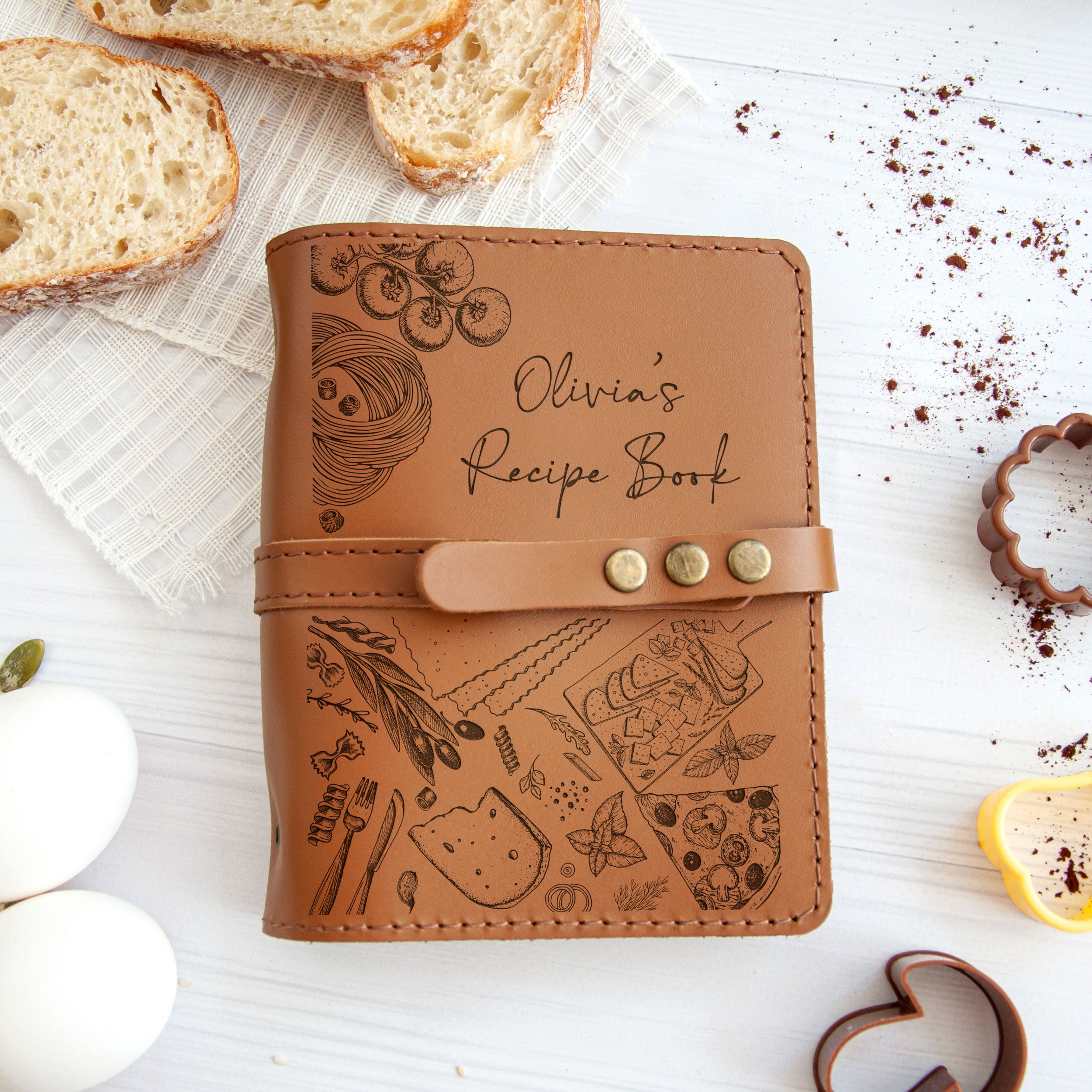 A personalized leather recipe book with various food items and utensils printed on it, along with a loaf of bread, a cookie cutter, and some crumbs on a white surface.