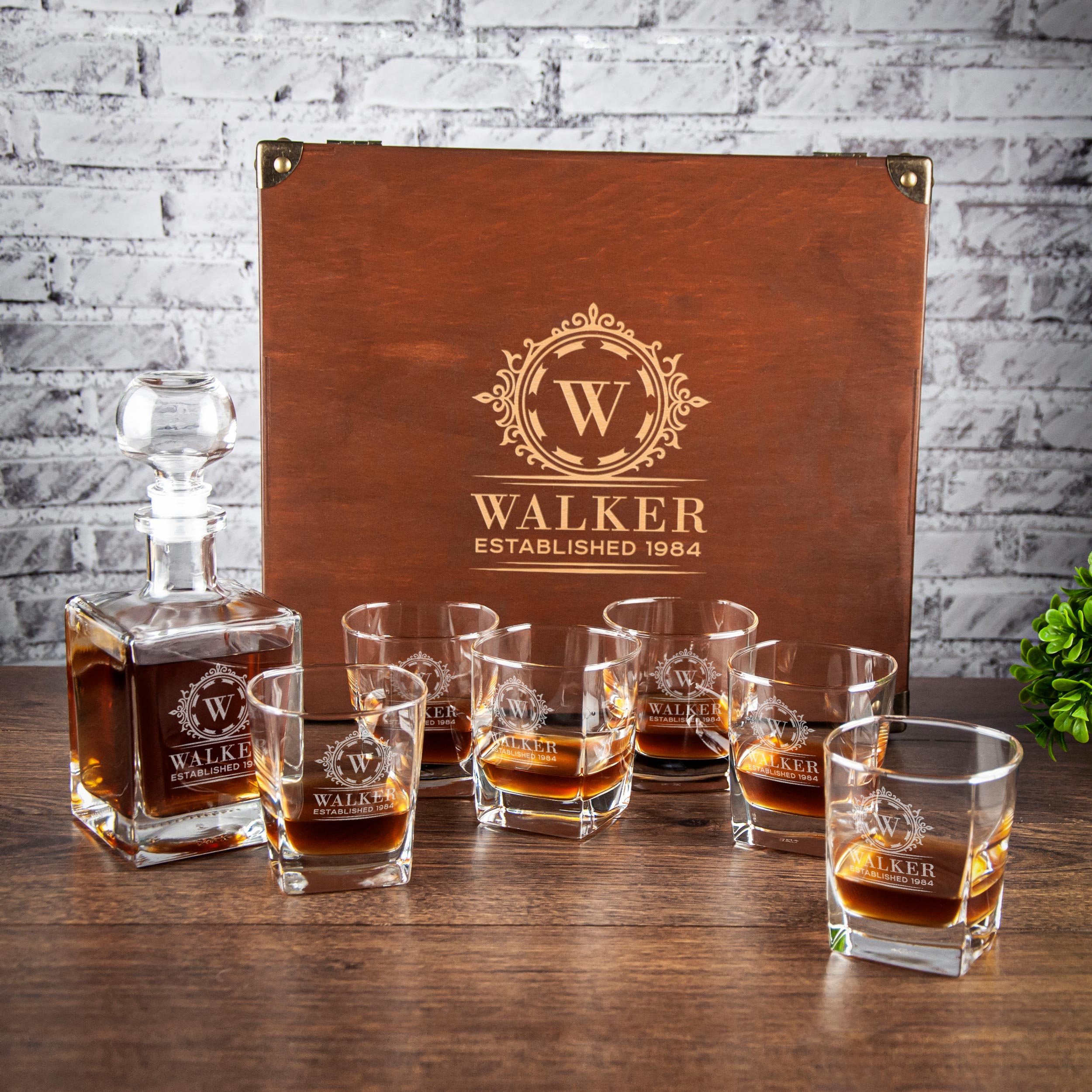 A set of six whiskey glasses and a wooden decanter with a gold logo, placed on a wooden table against a brick wall background.