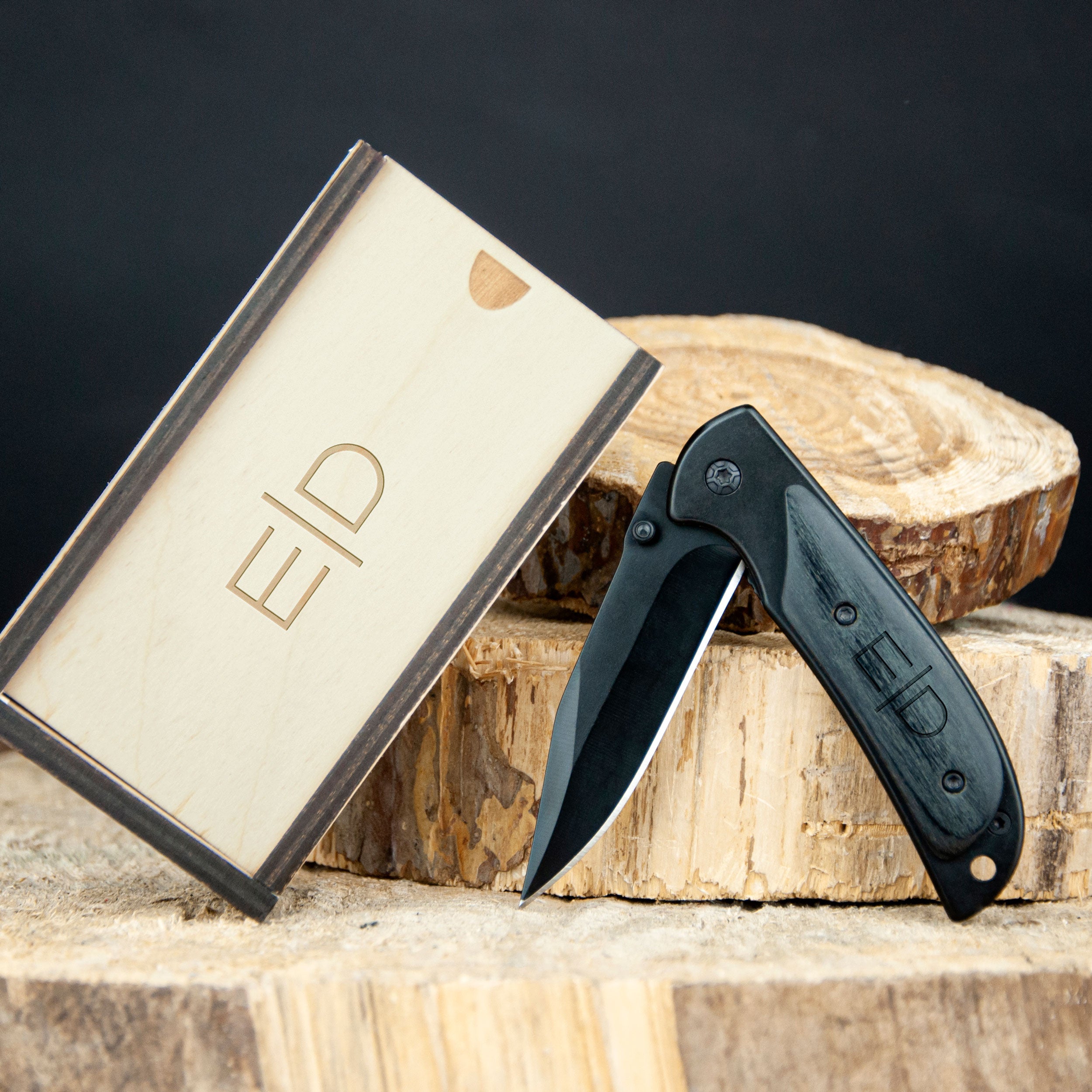 A black folding knife and a wooden box with the letters "E1D" on it are placed on a wooden surface.