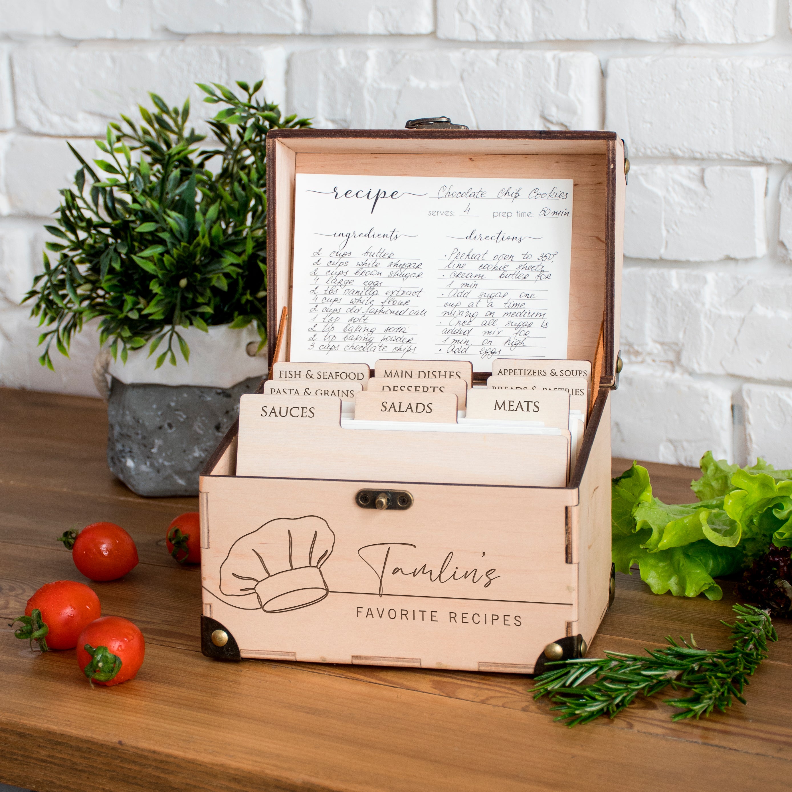 A wooden recipe box with a chef's hat on it, filled with recipe cards, sits on a wooden table next to a potted plant and some fresh vegetables.