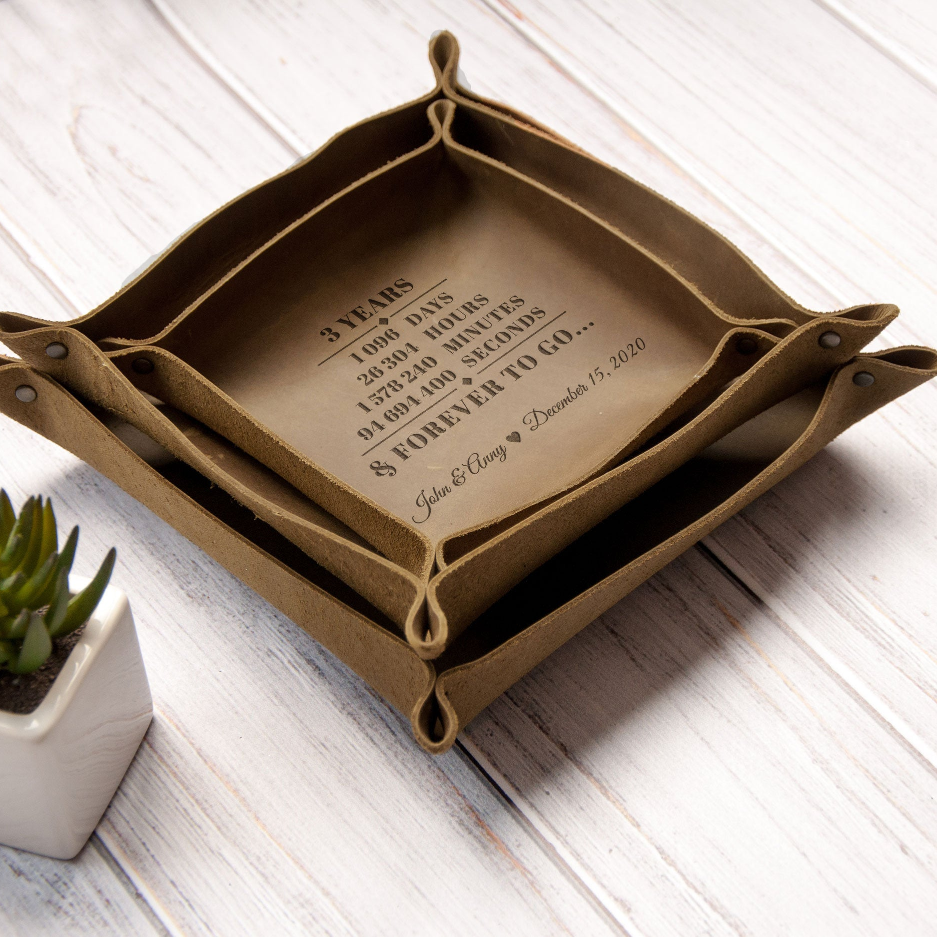 A brown leather tray with a message about a 3-year anniversary, placed on a white wooden surface next to a small potted plant.