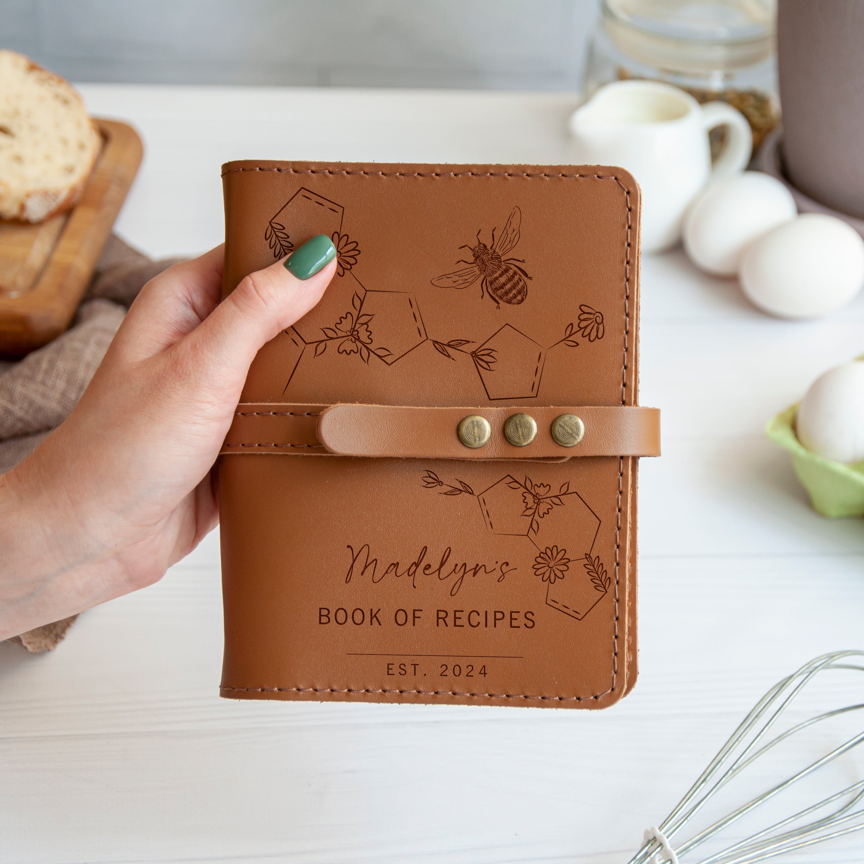 A hand holding a leather-bound book titled "Madelyn's Book of Recipes" with a bee and honeycomb design on the cover.