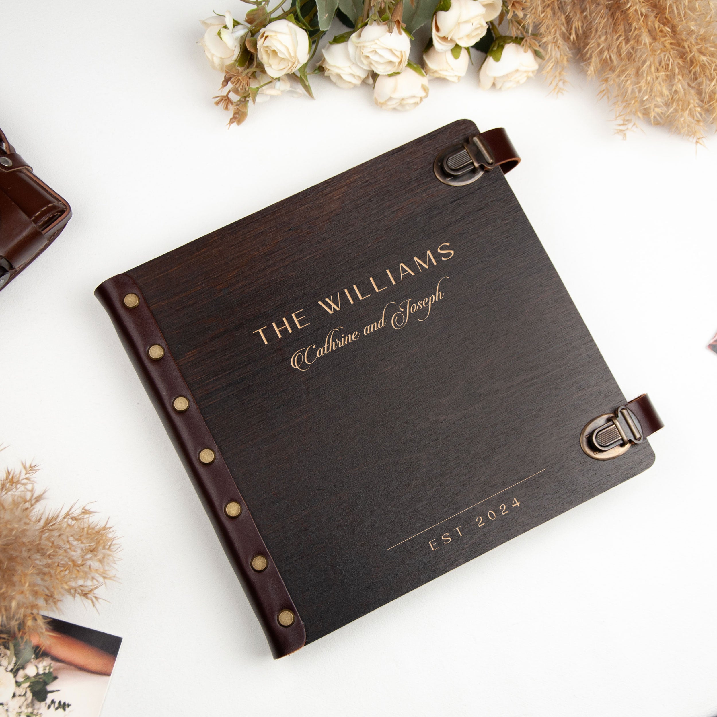 A dark brown leather-bound book titled "THE WILLIAMS" with the names "Cathrine and Joseph" printed on the cover, placed on a white surface alongside some flowers and a brown leather object.