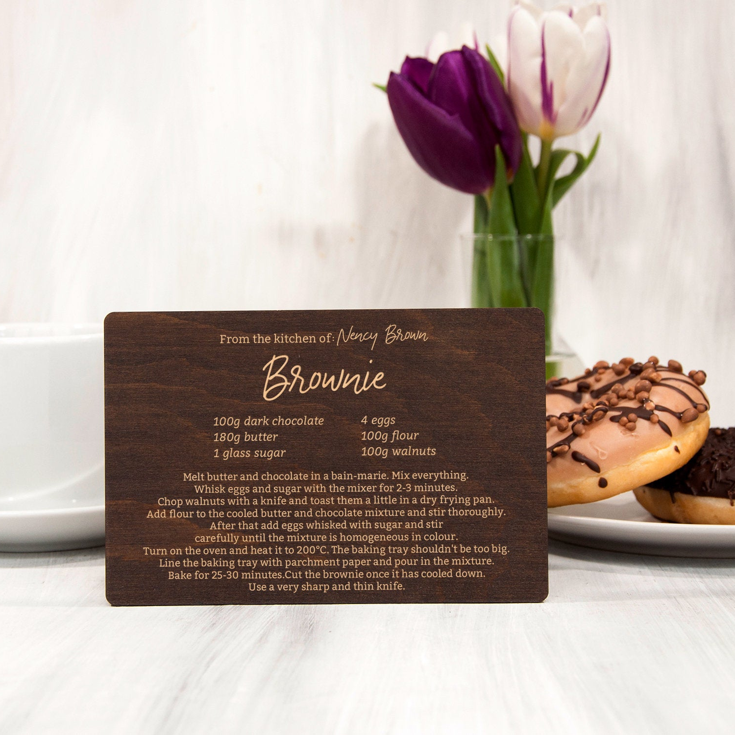 A wooden recipe card for a "Brownie" dessert, placed on a table with a vase of purple and white tulips in the background.