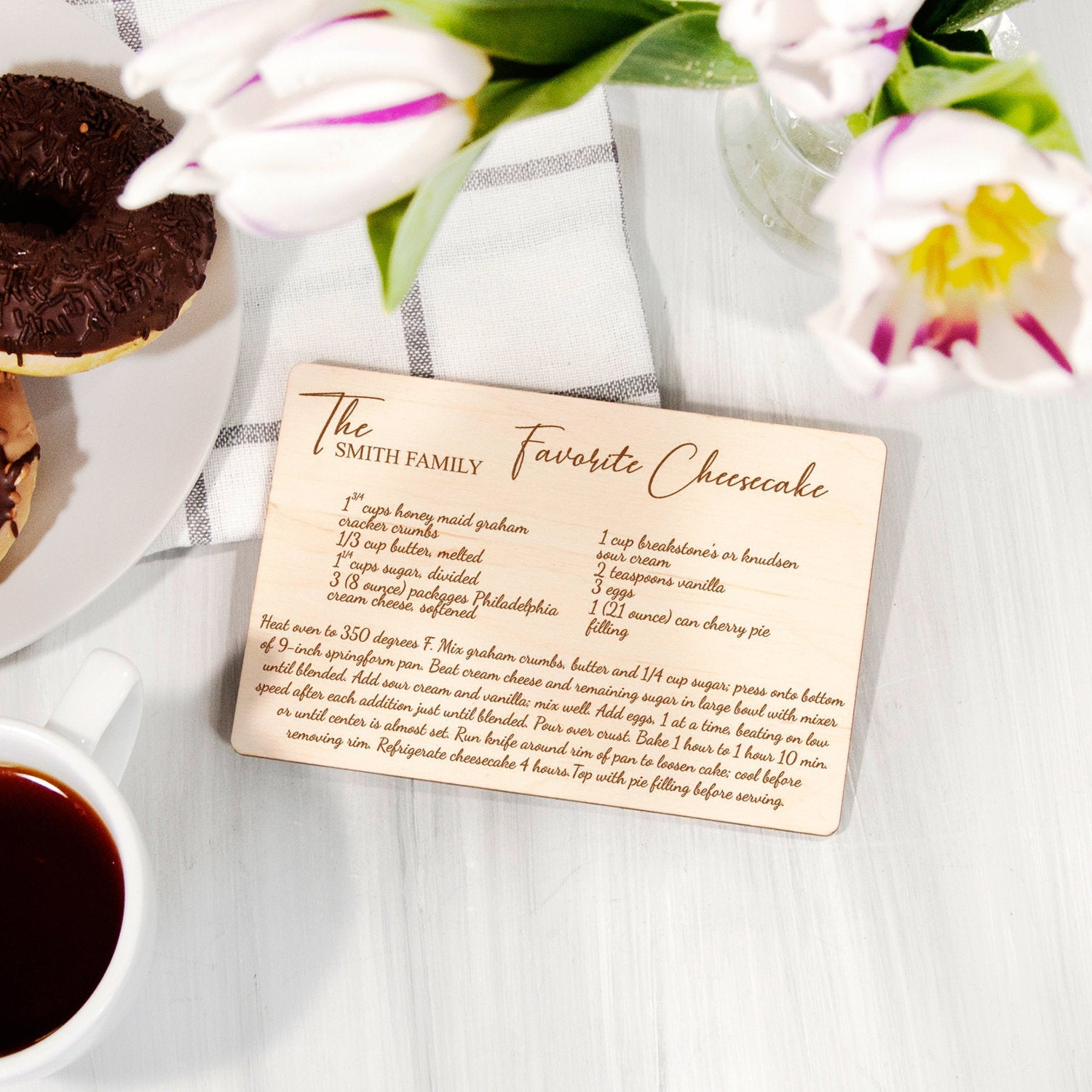A wooden recipe card titled "The Smith Family Favorite Cheesecake" is placed on a table next to a plate of chocolate-covered doughnuts and a vase of white and purple flowers.