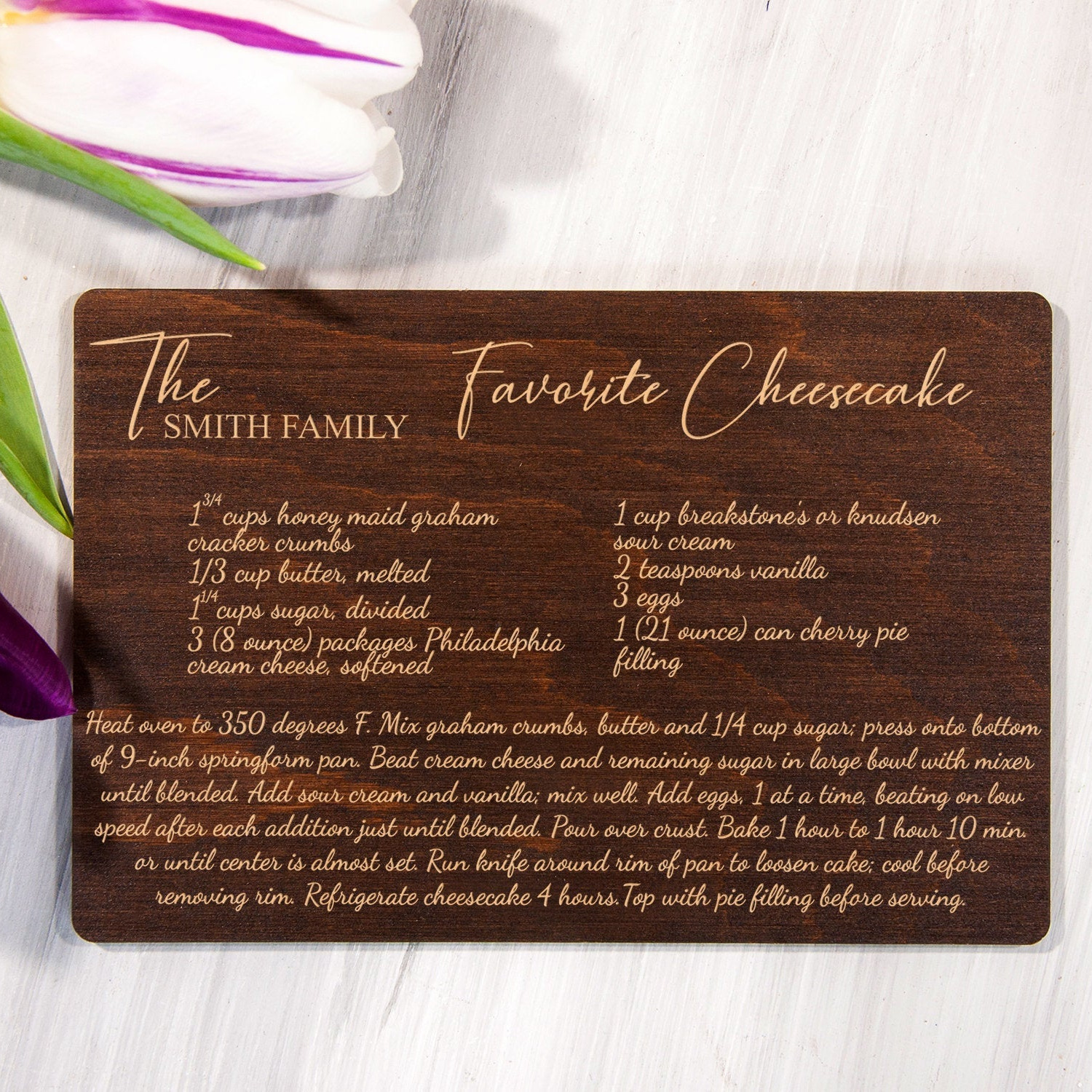 A wooden recipe card titled "The Smith Family Favorite Cheesecake" is displayed on a white surface, accompanied by a purple flower.